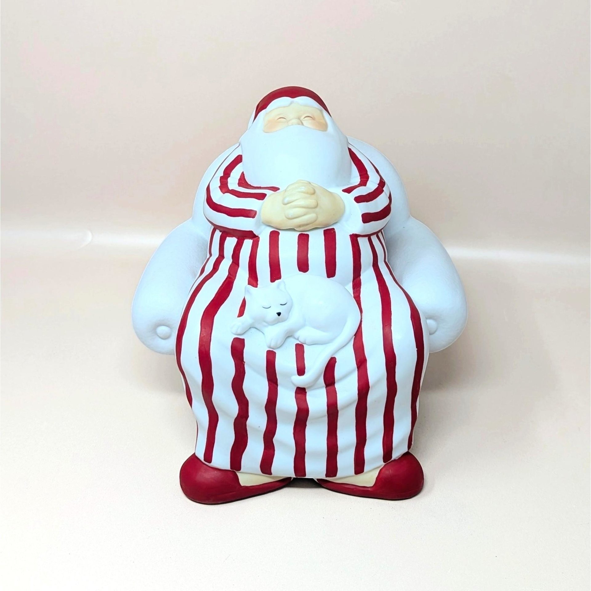 A vintage Department 56 Winter Silhouettes "Cat Nap" Santa figurine on a soft pink background. The matte white porcelain sculpture shows Santa sleeping soundly in a large armchair wearing red-striped pajamas with a white cat napping on his lap.