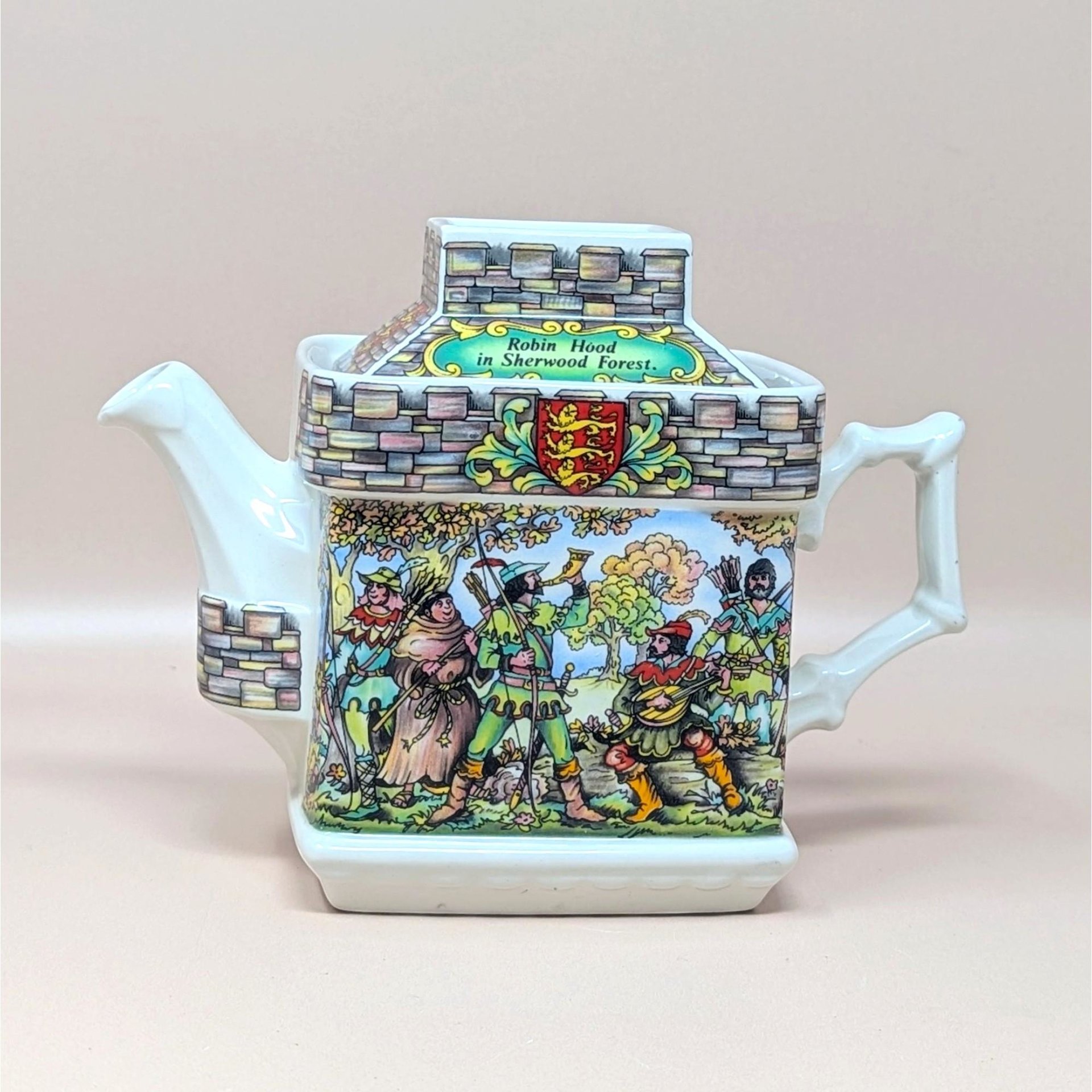 A vintage Sadler porcelain castle themed teapot on a soft pink background. The square cream colored teapot features a detailed double sided illustration of the Sheriff of Nottingham at his table and Robin Hood in Sherwood Forest with stone walls.