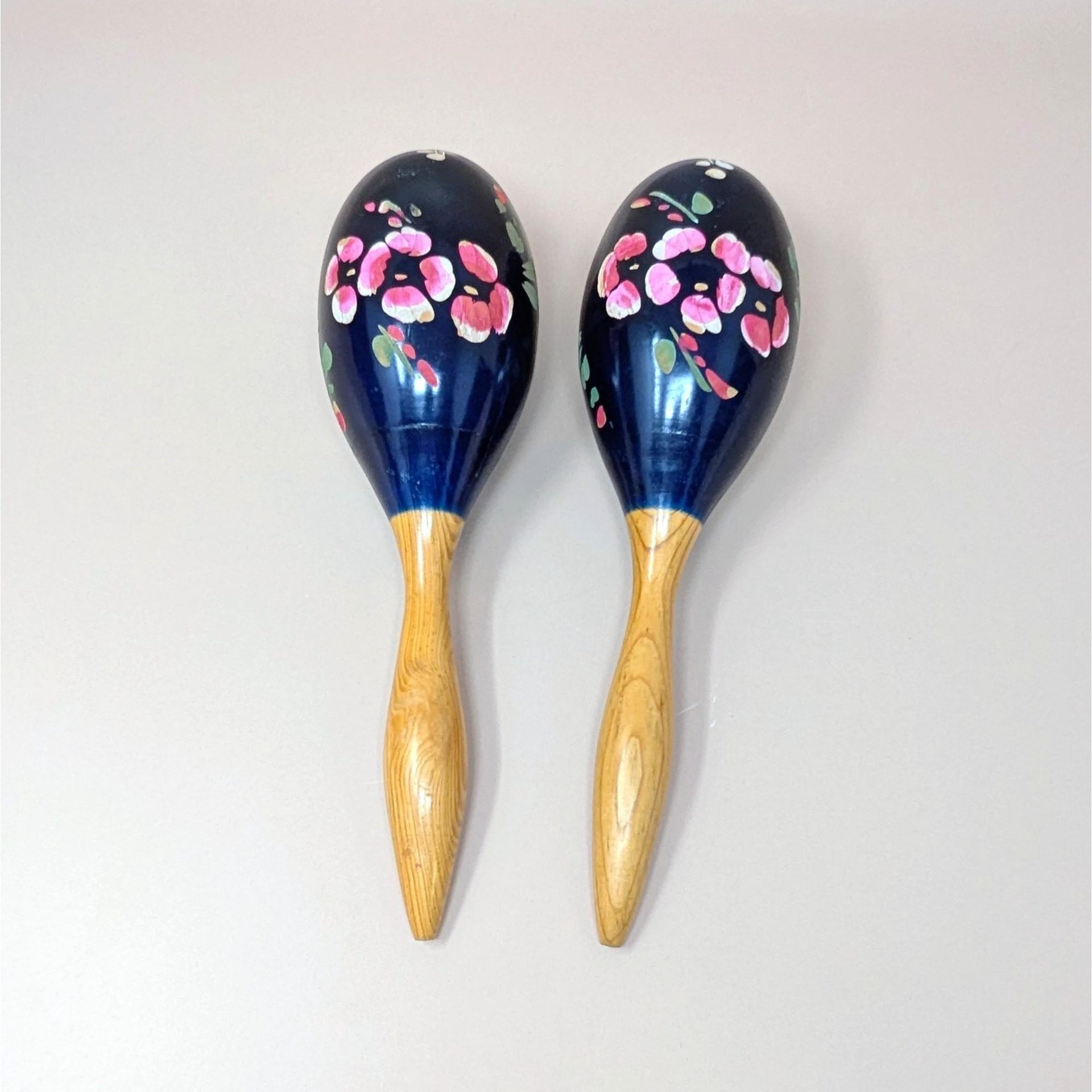 A pair of vintage hand painted wooden maracas on a soft pink background. The musical instruments feature sleek natural wood grain handles and glossy dark blue bulbs decorated with hand painted bright pink and red flowers.