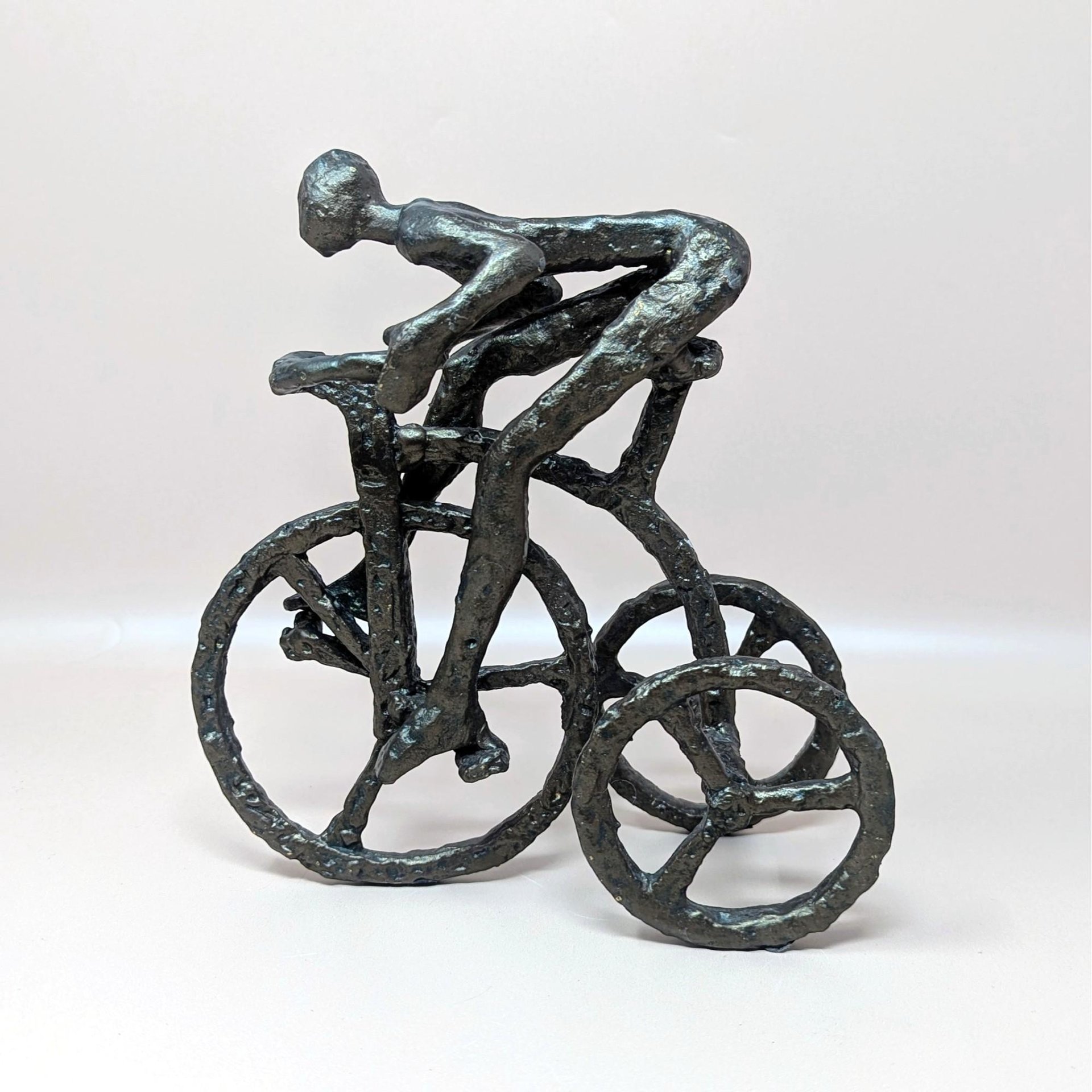 A vintage brutalist cast iron sculpture of a man riding a tricycle on a soft pink background. The dark, heavily textured metal figurine depicts a stylized, faceless human figure leaning forward in a racing position over three spoked wheels.