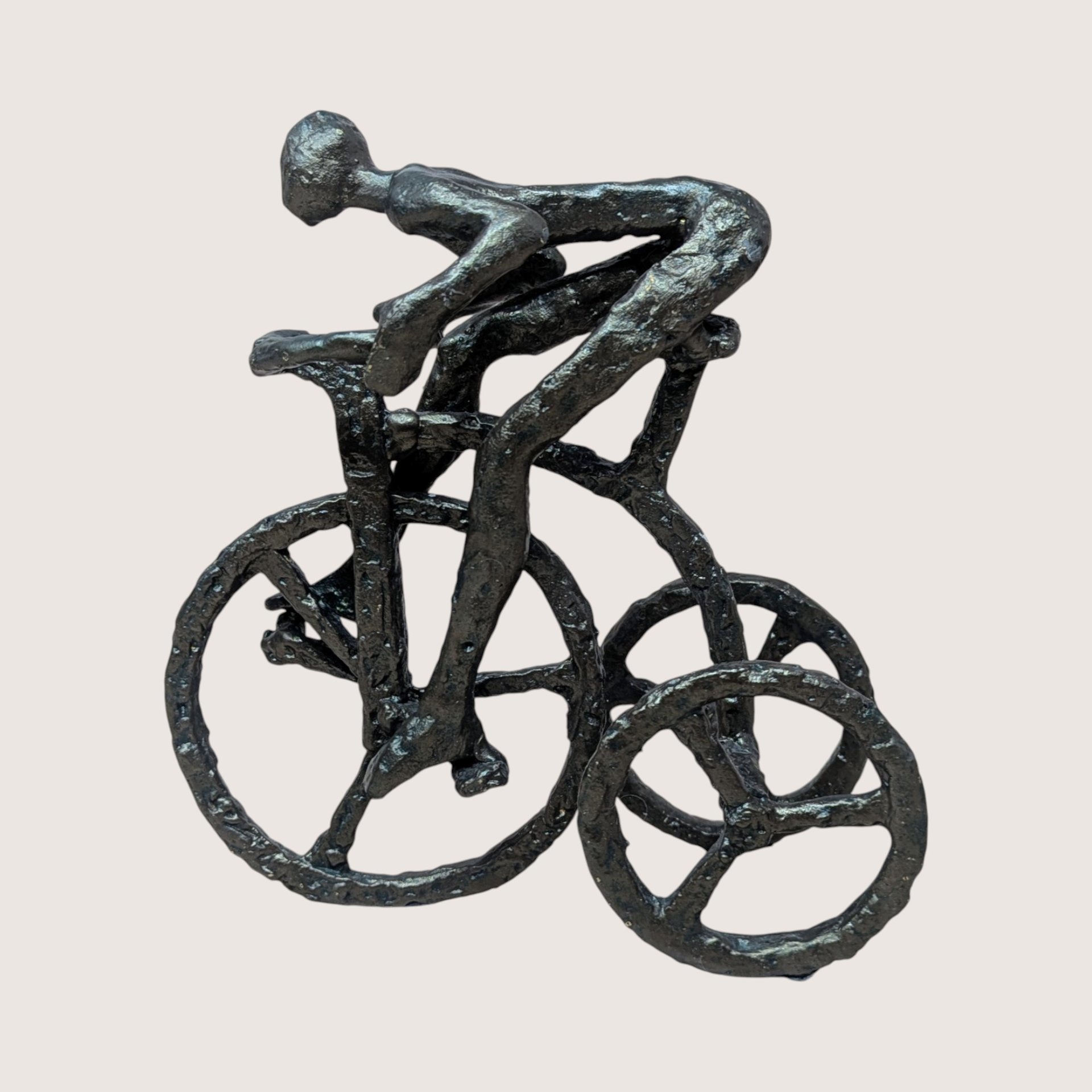 A vintage brutalist cast iron sculpture of a man riding a tricycle on a soft pink background. The dark, heavily textured metal figurine depicts a stylized, faceless human figure leaning forward in a racing position over three spoked wheels.