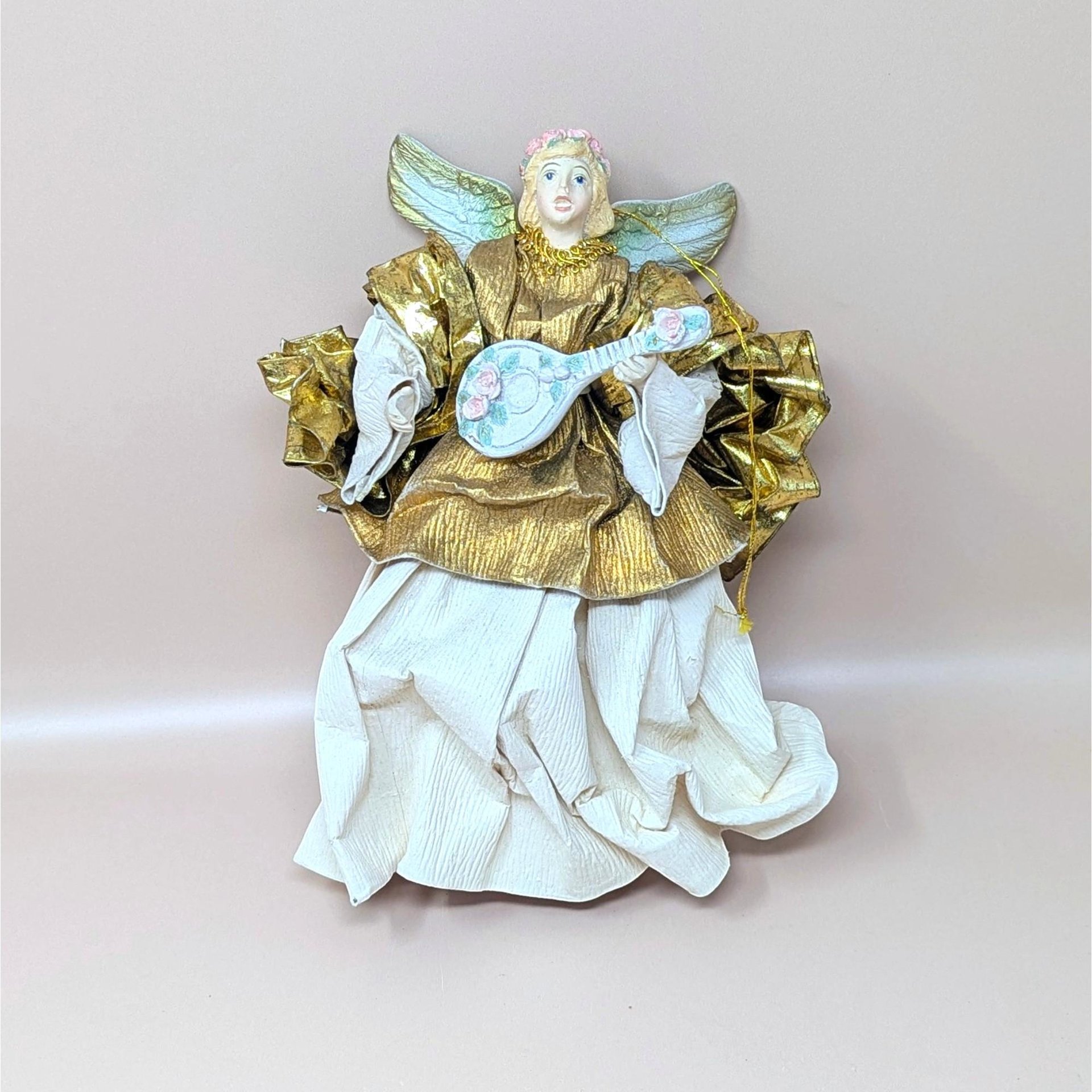 A vintage paper mache and fabric angel tree ornament on a soft pink background. The nine inch tall angel features a metallic gold upper gown, stiffened cream fabric crinkle skirt, soft gold wings, and holds a small blue stringed lute.