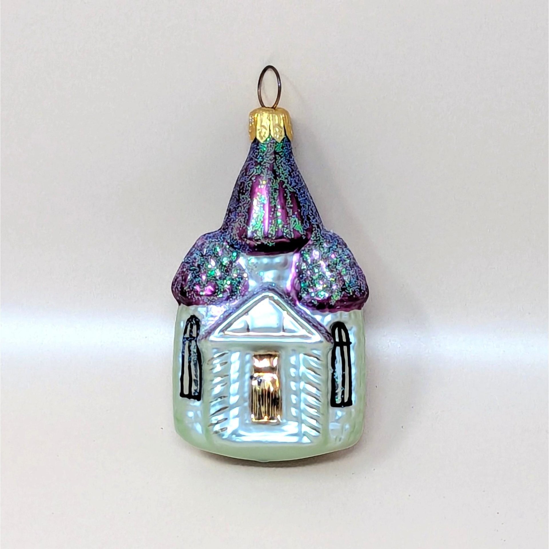 A vintage Christopher Radko blown glass church ornament with its box on a soft pink background. The fine mercury glass cathedral features iridescent pale green walls, hand painted black windows, and shimmering purple steeples covered in sparkling glitter.