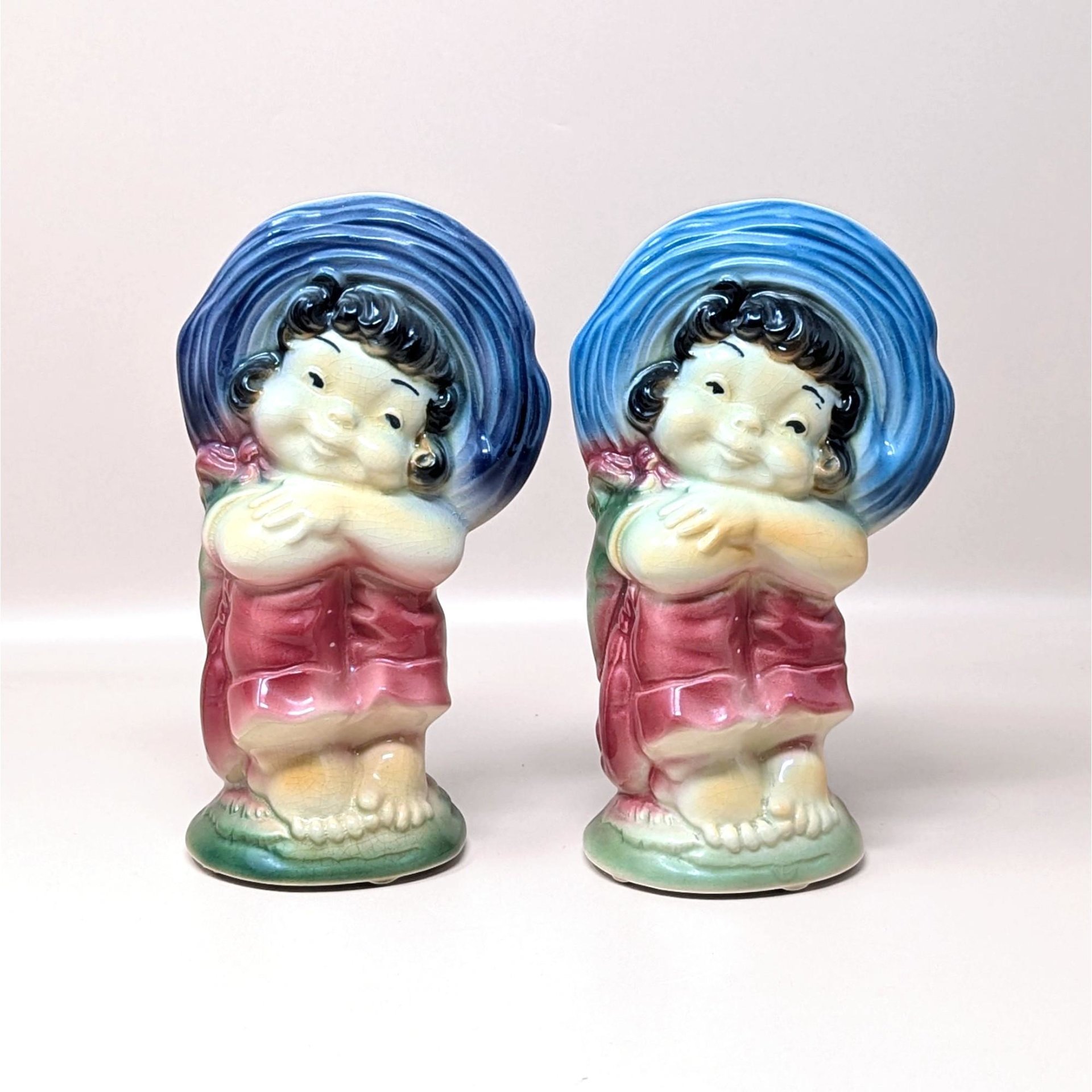A pair of vintage Royal Copley ceramic planters on a soft pink background. Made in the 1950s, each matching vase features a charming barefoot girl wearing a massive blue sunbonnet and a pink dress with a glossy, airbrushed finish.