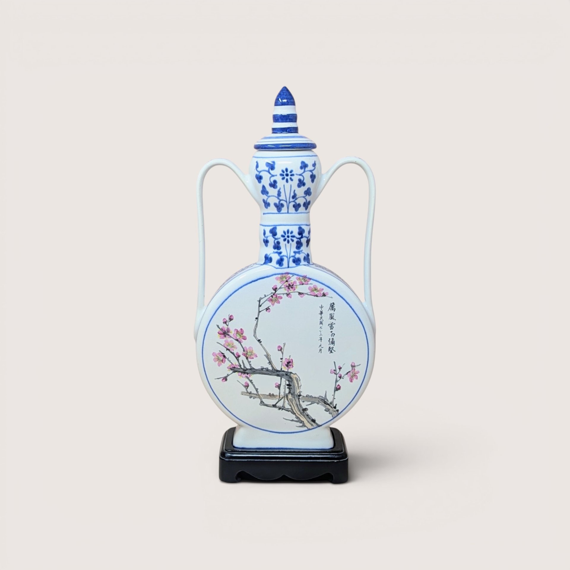 A large blue and white porcelain moon flask with pink plum blossoms and Chinese calligraphy on a wooden stand against a plain background