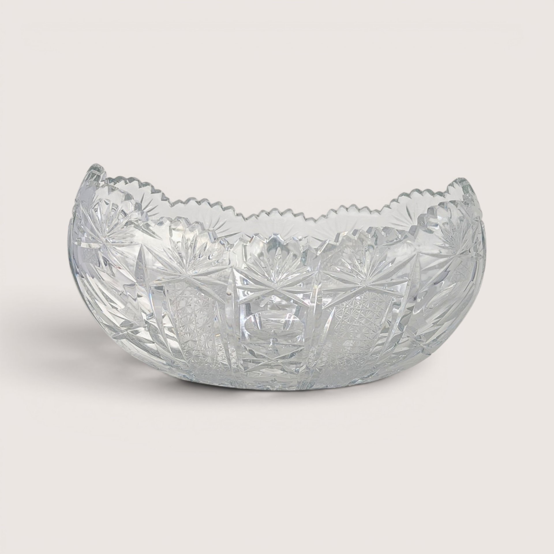 An oval clear cut glass bowl with sharp geometric starburst patterns and a sawtooth rim on a plain background