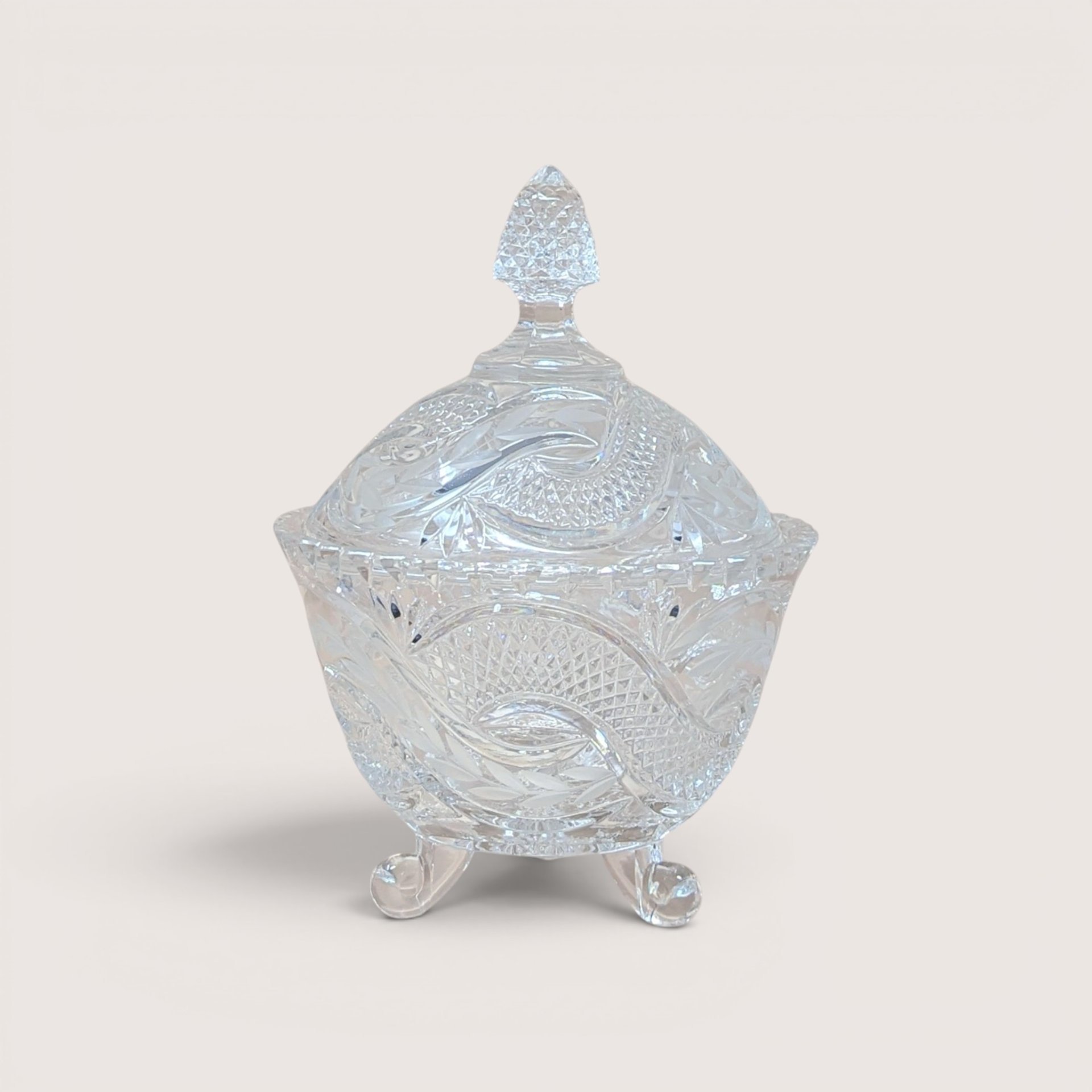 A vintage clear crystal candy dish with a matching lid and three scroll feet on a plain background