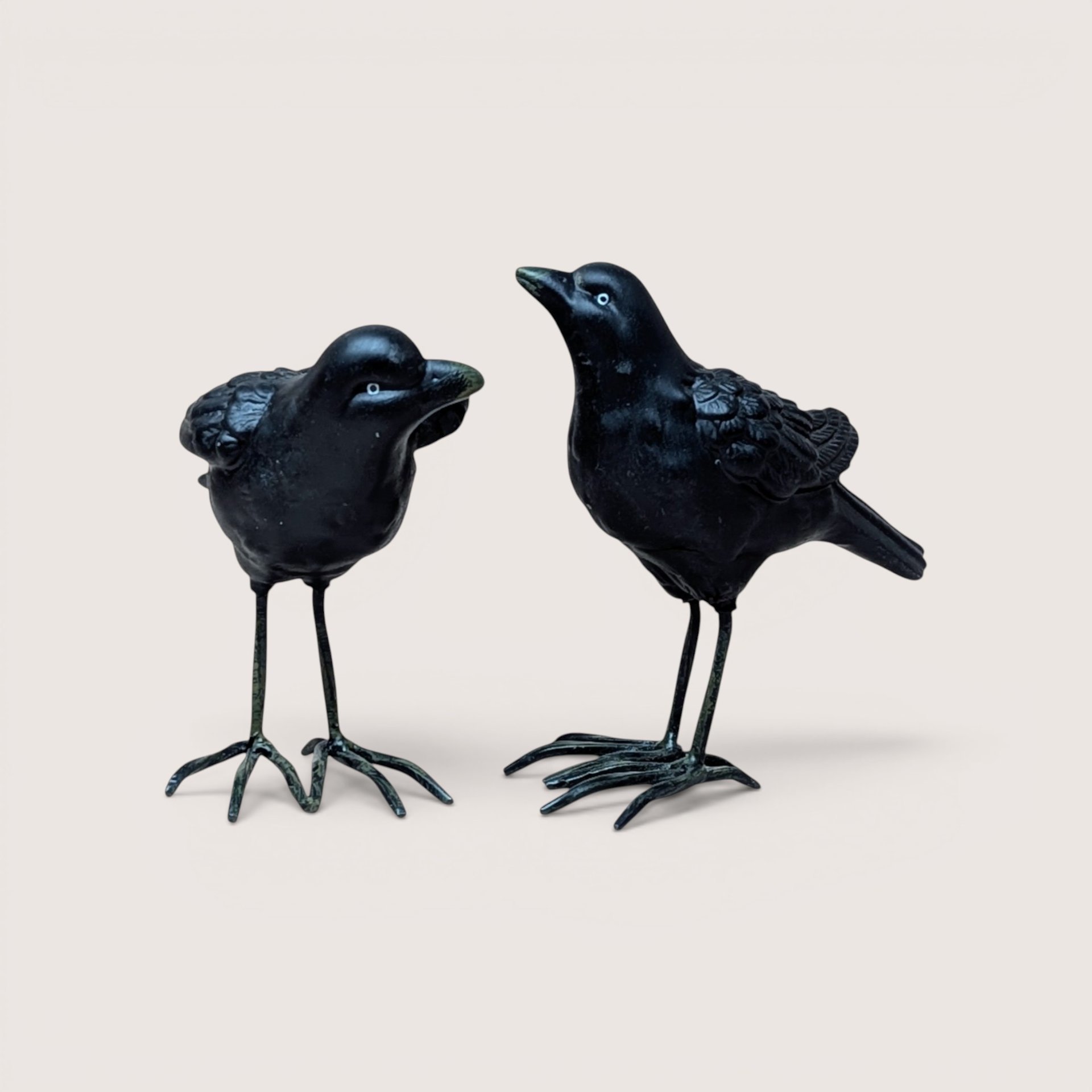 Two black ceramic crow figurines with long metal legs standing side by side on a plain background