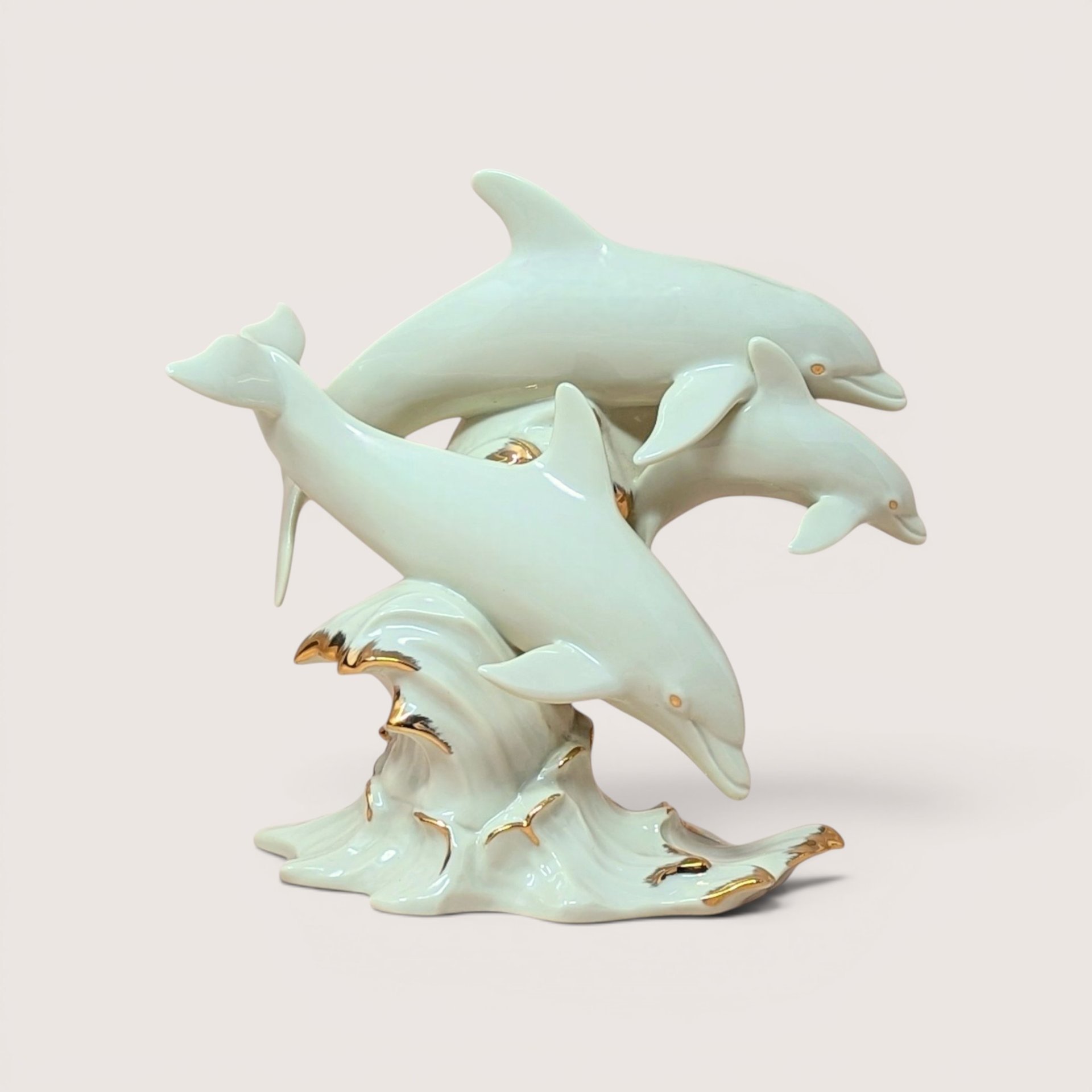 Three ivory porcelain dolphins leaping from a gold trimmed ocean wave on a solid cream colored background