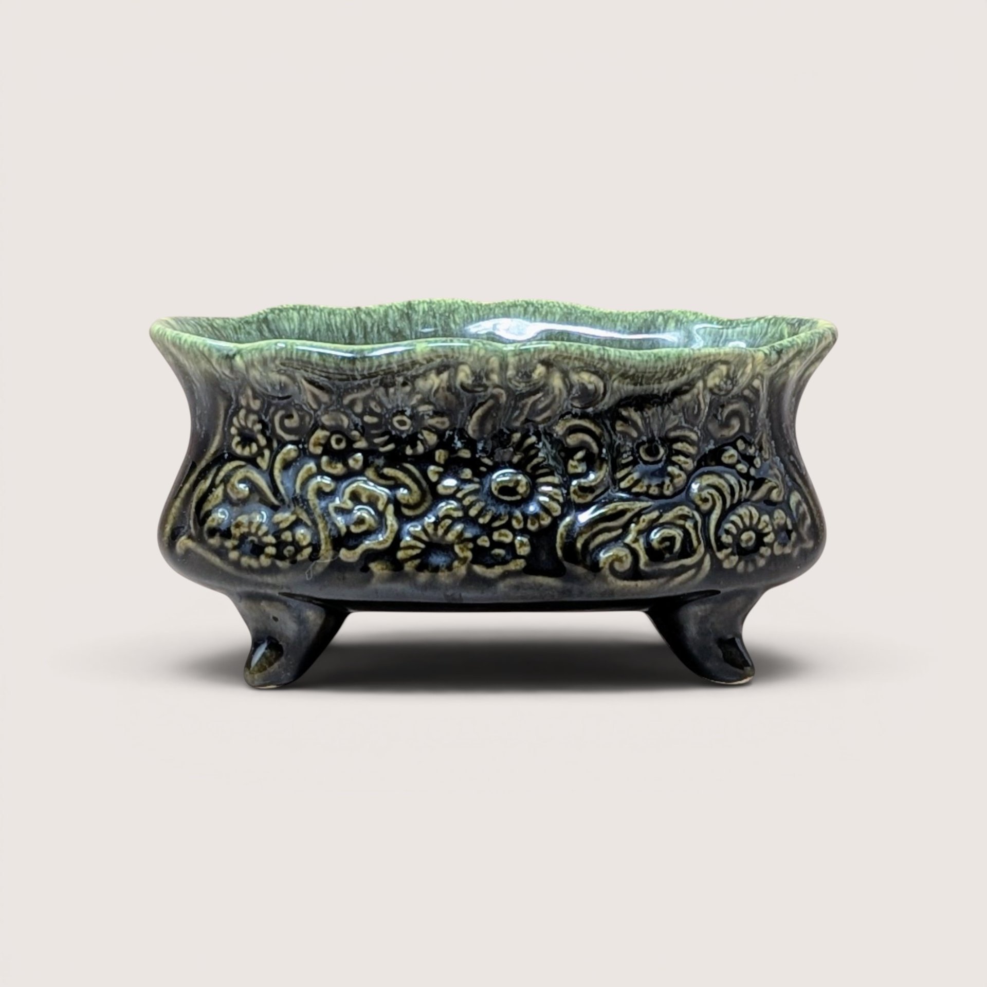 A vintage footed green ceramic planter with detailed embossed flowers resting on a plain beige background