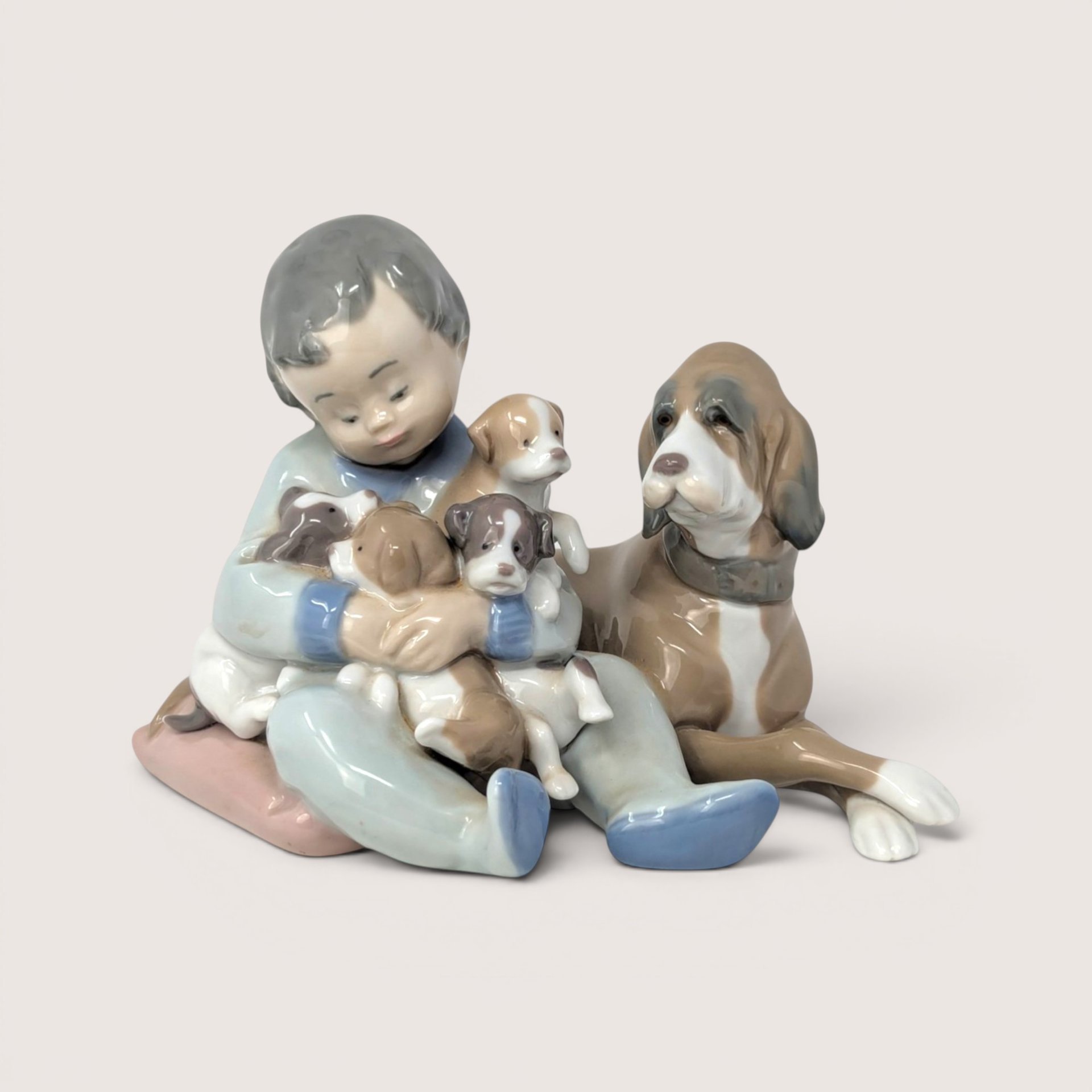 A vintage porcelain figurine of a young boy sitting on a pink cushion holding four puppies next to a large dog
