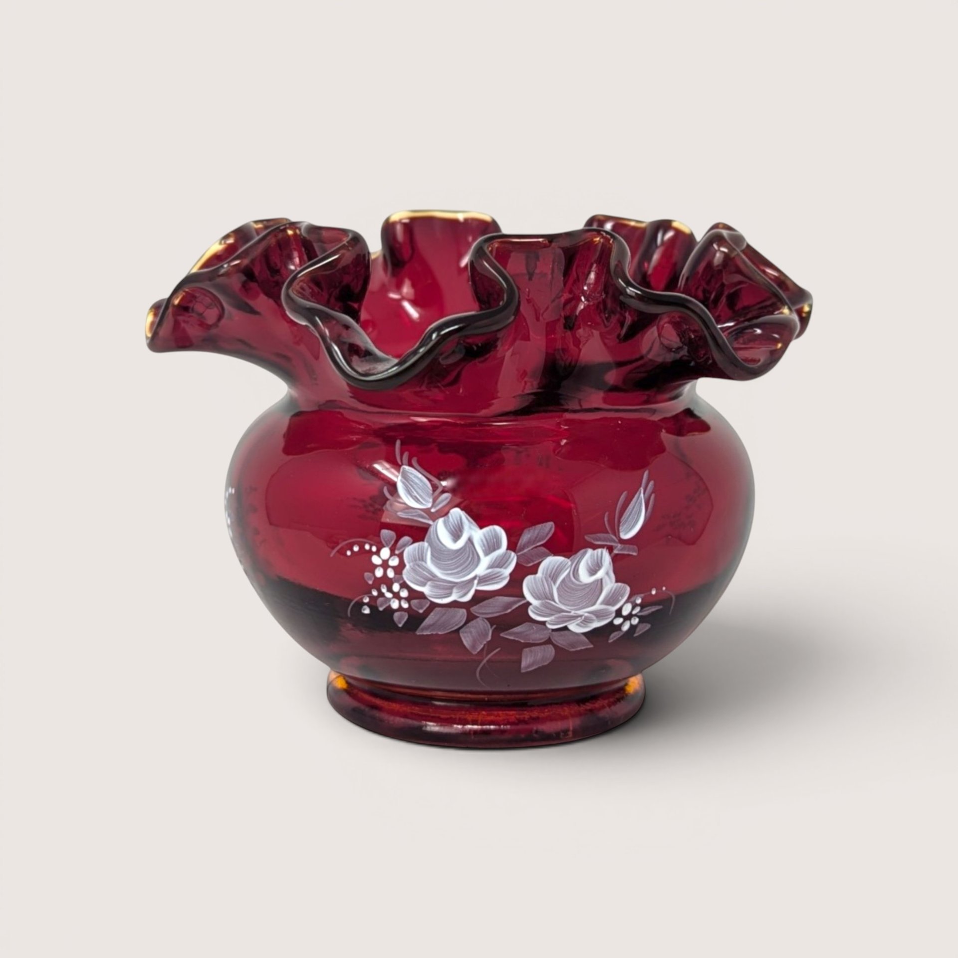 A vintage ruby red glass vase with a deeply ruffled rim and hand painted white roses on a plain background