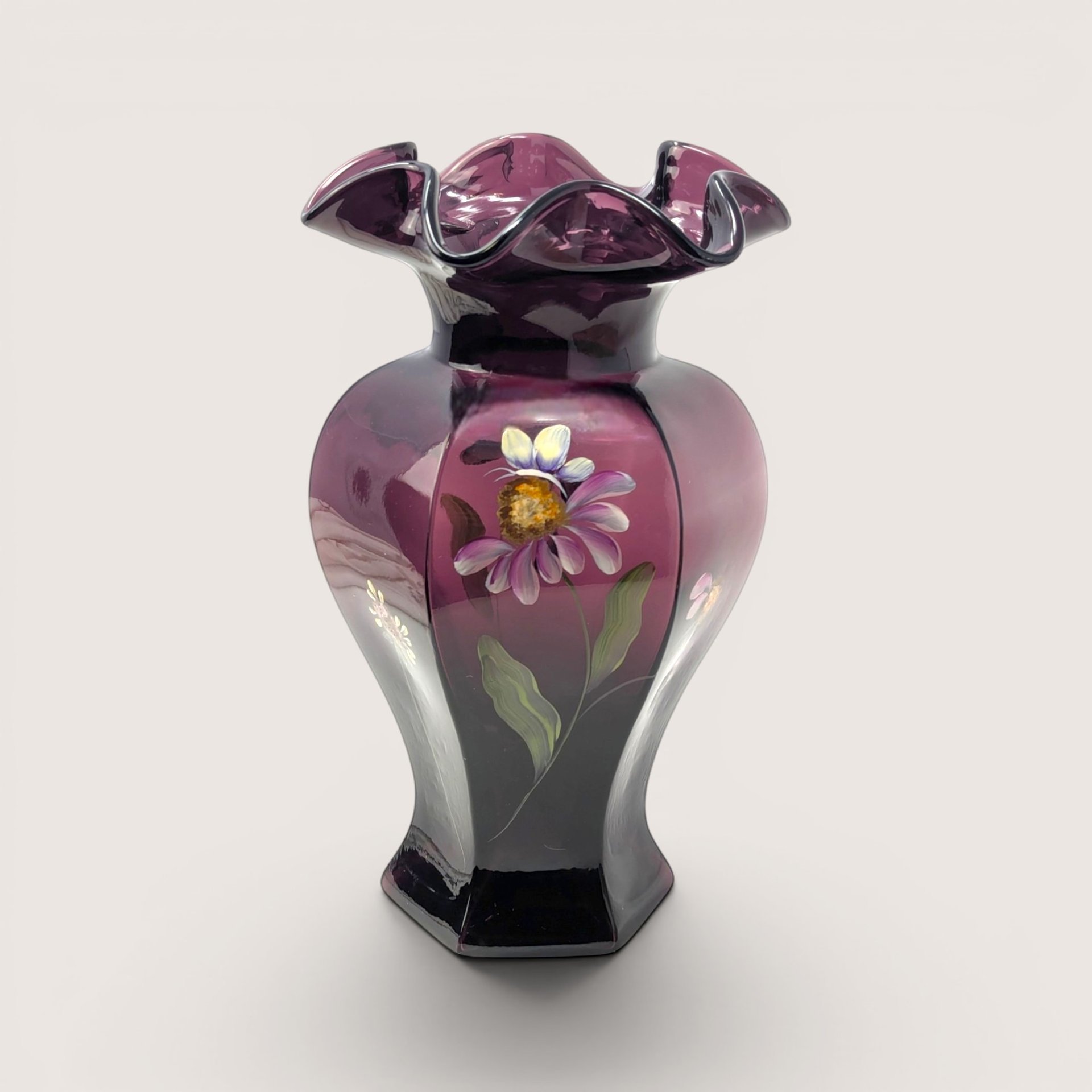 A dark purple aubergine glass vase with a ruffled rim and hand painted purple flower on a light beige backdrop