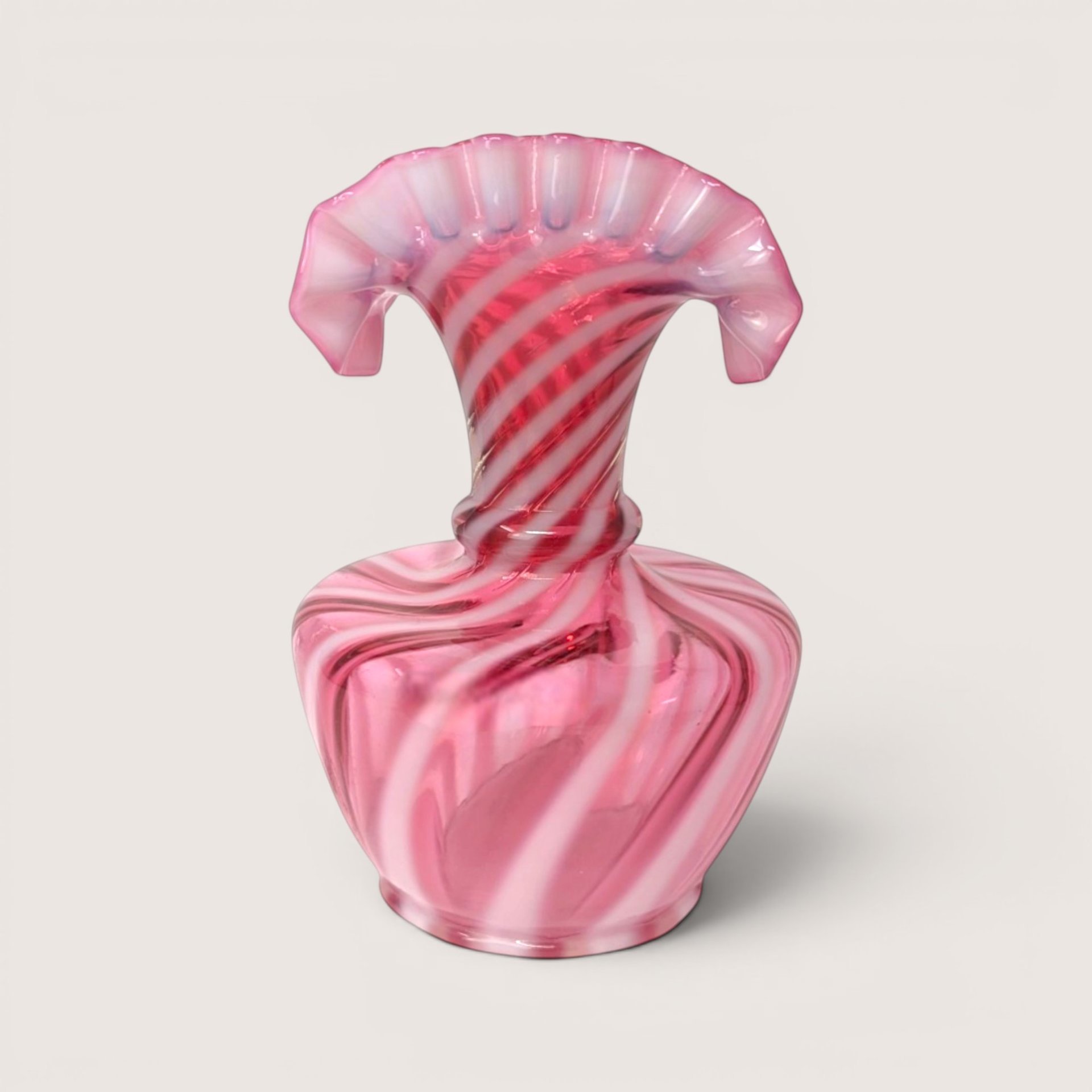 A pink and white spiral swirl opalescent glass fan top vase with a wide flattened ruffled rim on a beige backdropA pink and white spiral swirl opalescent glass jack in the pulpit vase on a light beige background