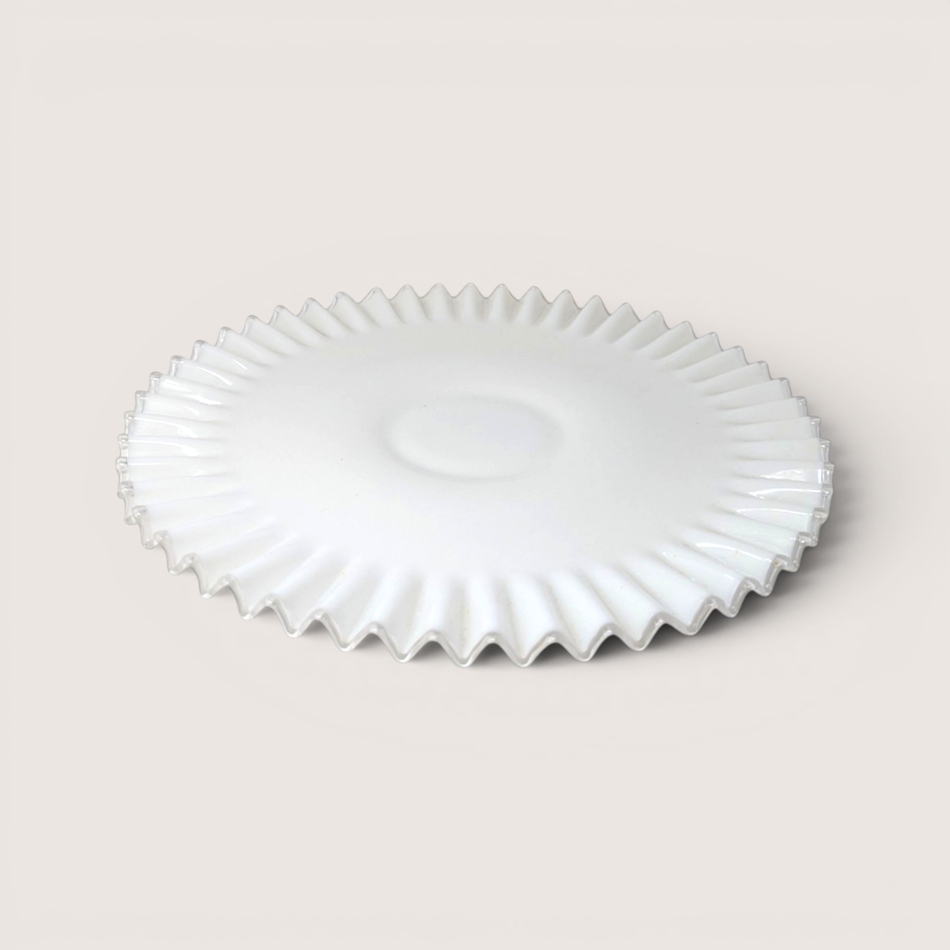 A round white milk glass cake salver with a clear ruffled edge on a light beige backdrop