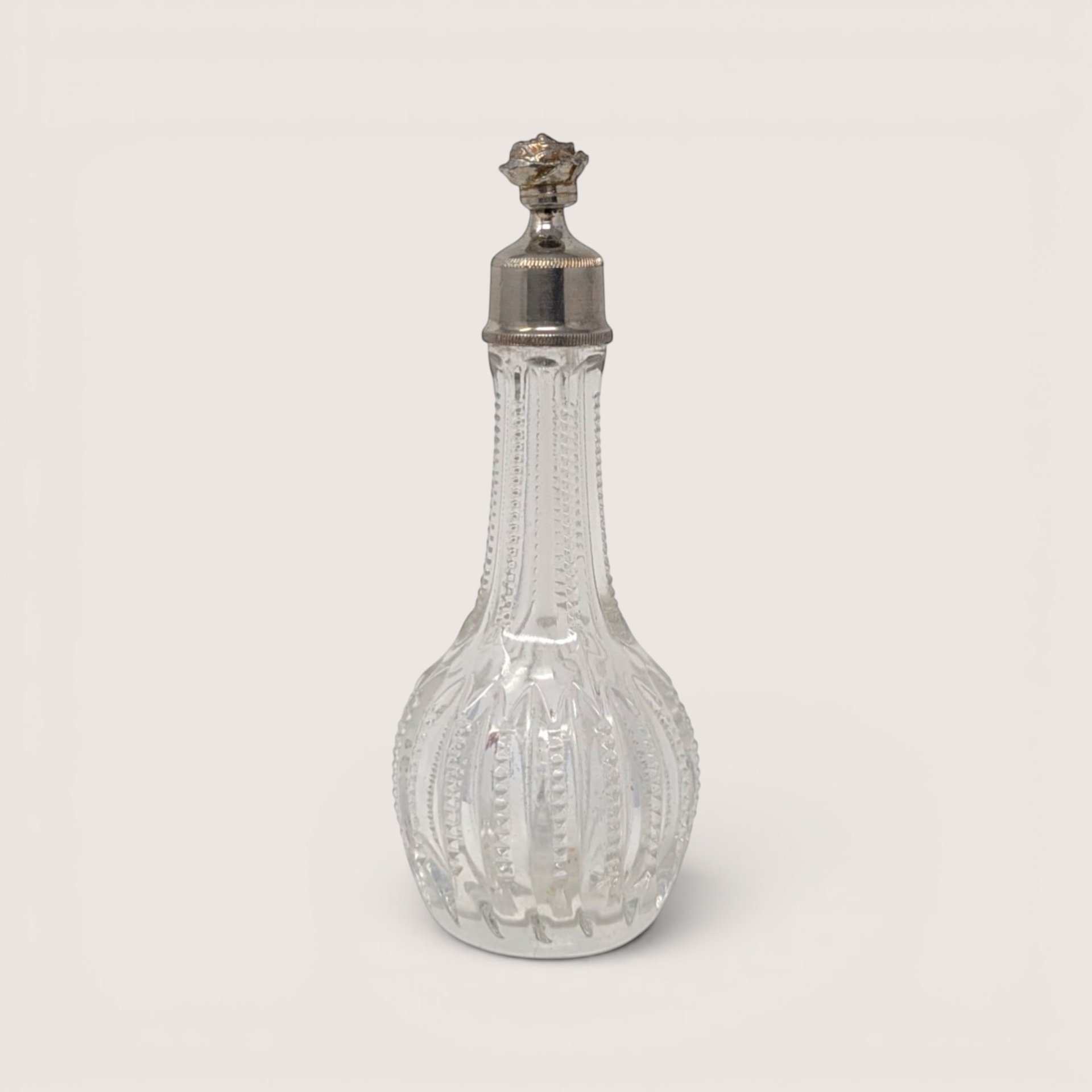 Antique ribbed clear glass perfume bottle with an ornate silver tone floral cap on a neutral background.