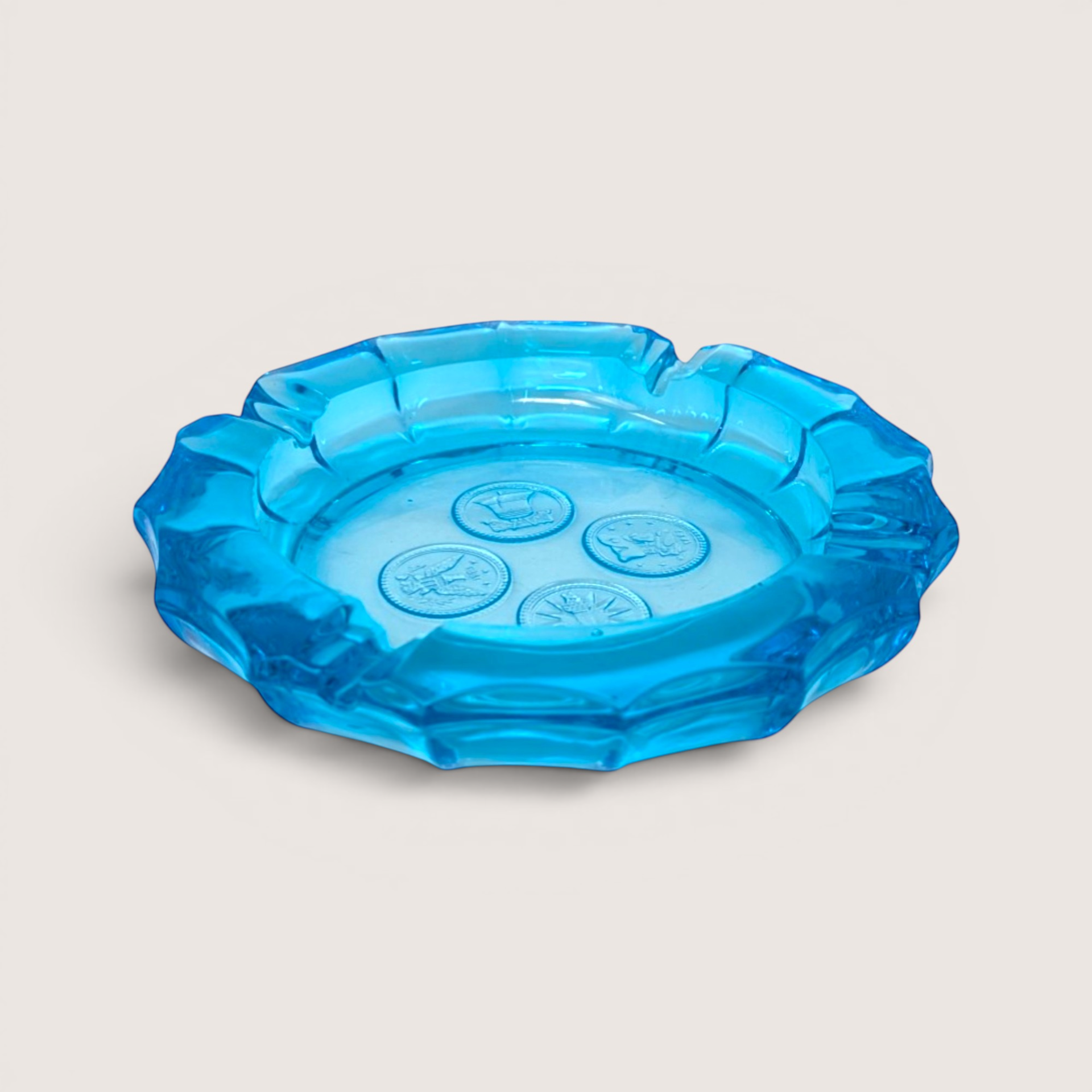 Vibrant blue glass ashtray featuring four embossed coin medallions in the center on a solid background.