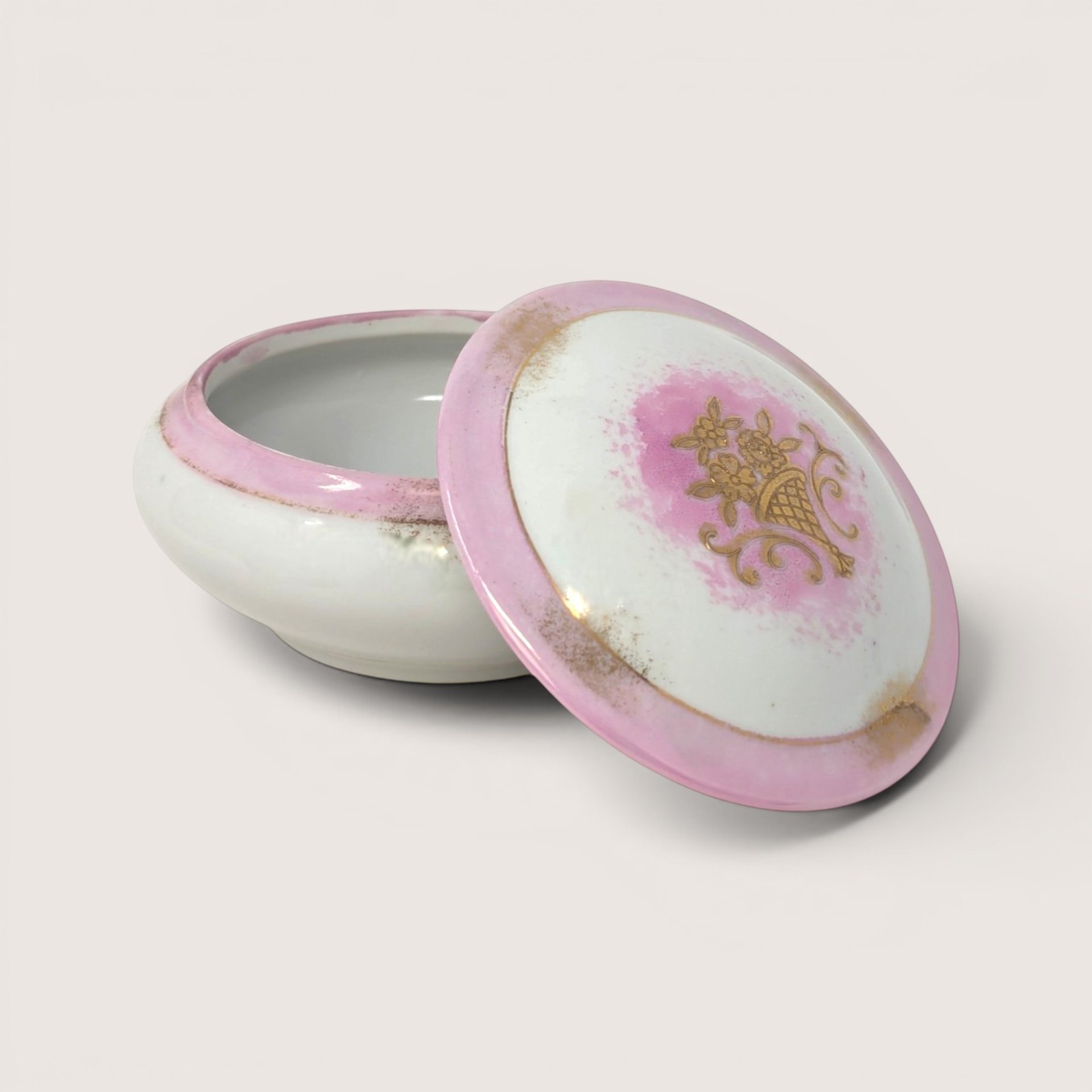 Round white porcelain trinket box with an open lid featuring a pink rim and a gold floral basket on a solid background.