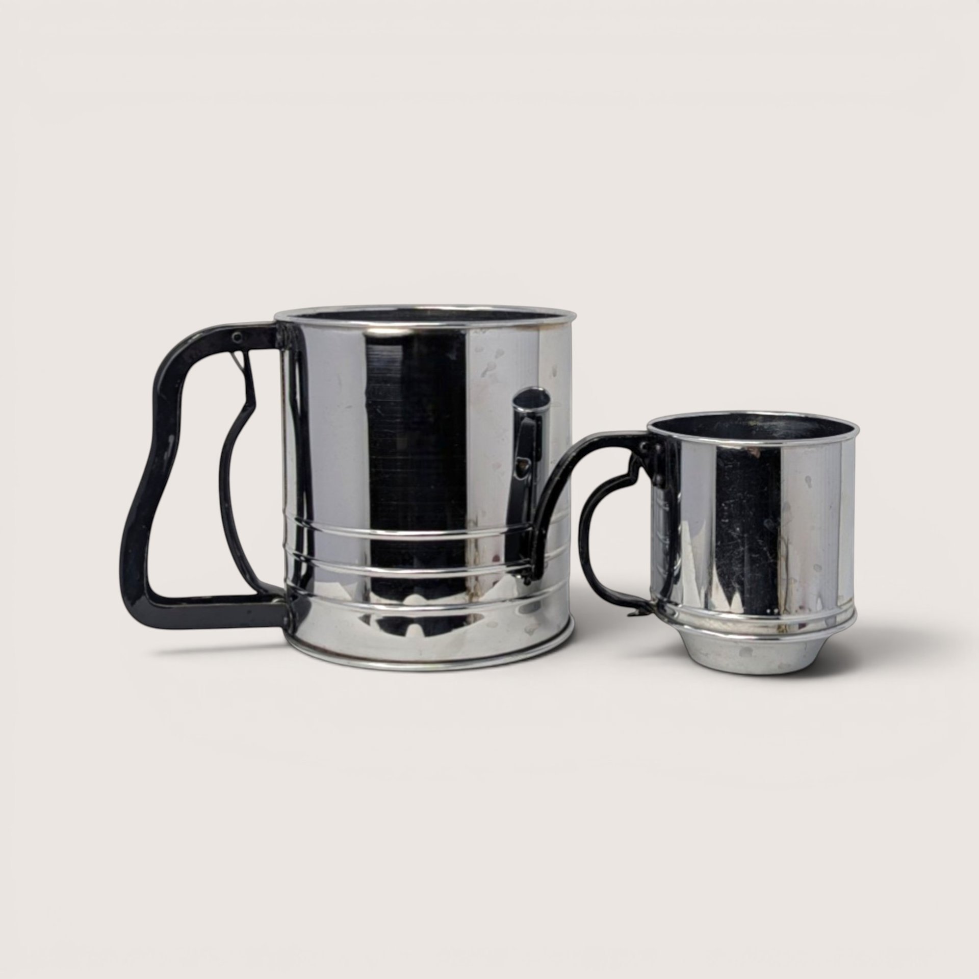 Two stainless steel flour sifters in large and small sizes with black handles on a solid background.