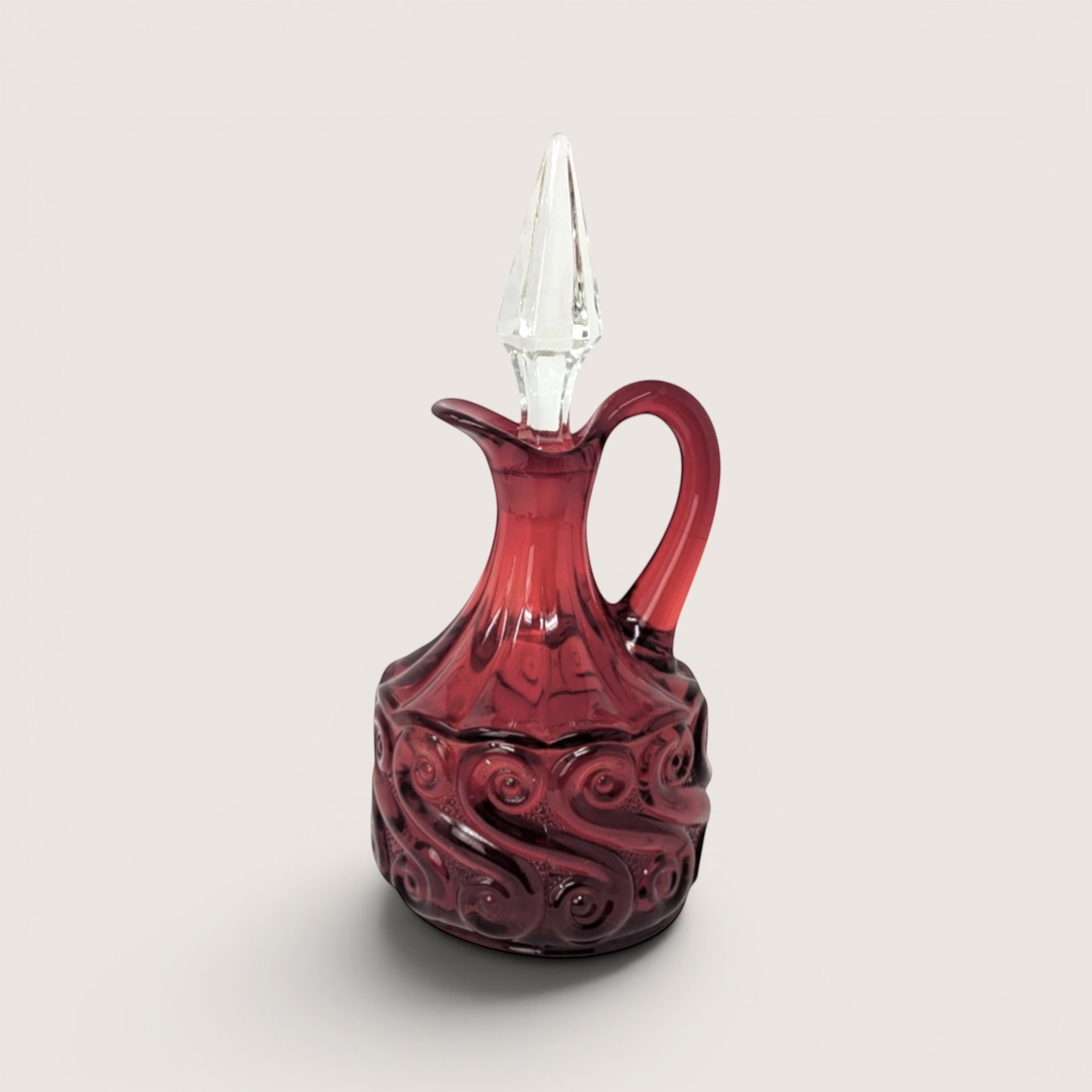 Ruby red pressed glass cruet with an embossed scroll pattern and a clear faceted crystal stopper on a solid background.