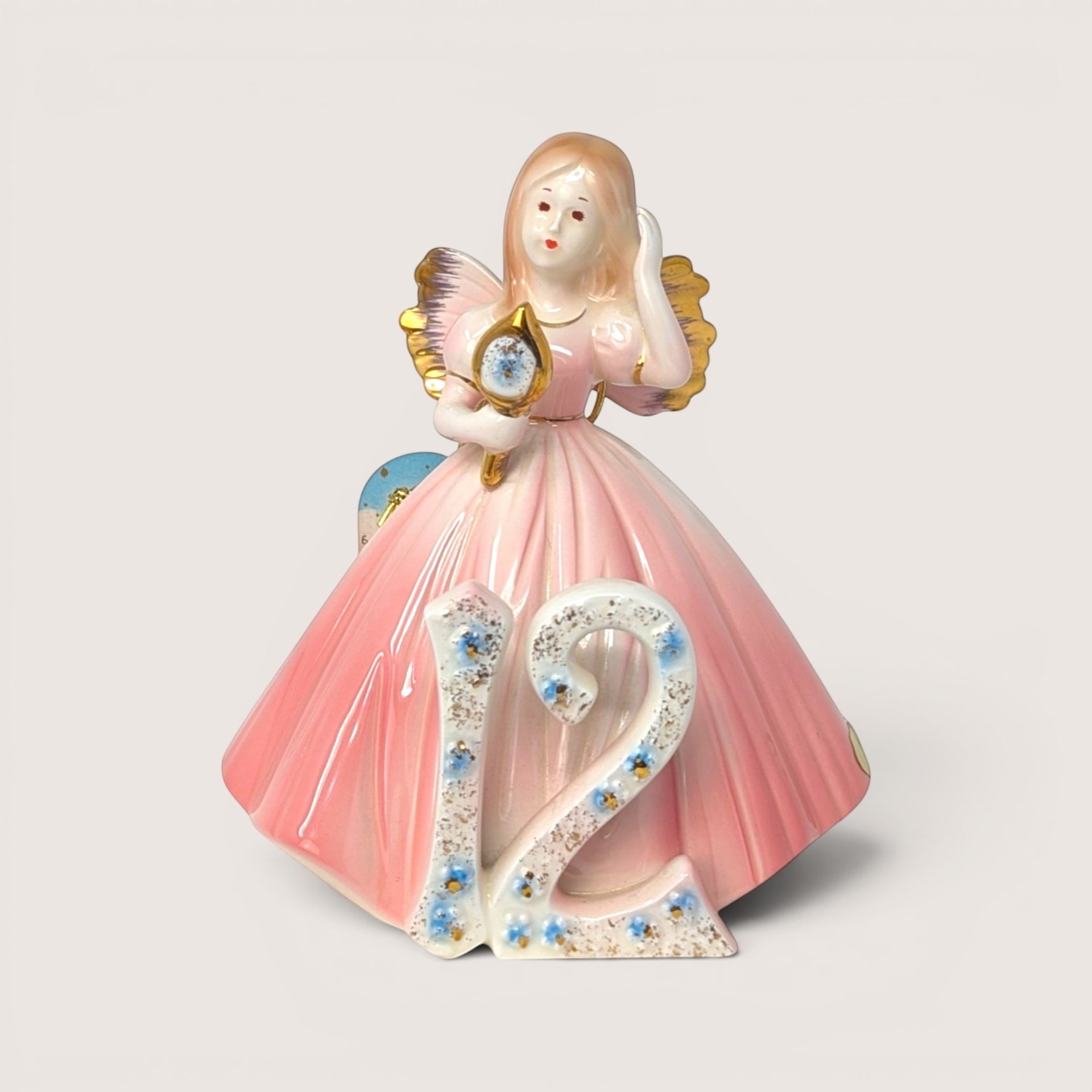 Porcelain birthday angel figurine in a pink gown holding a hand mirror behind a floral number 12 on a solid background.