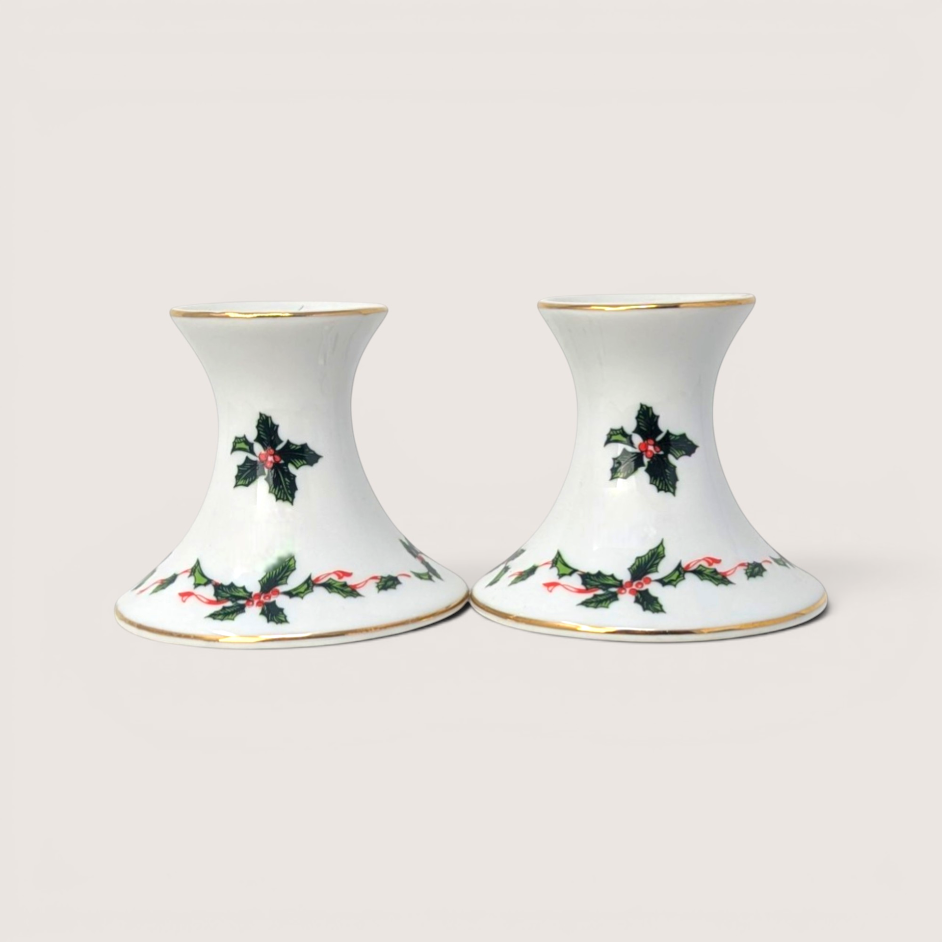 Pair of white ceramic candlestick holders decorated with green holly leaves, red berries, and gold trim.