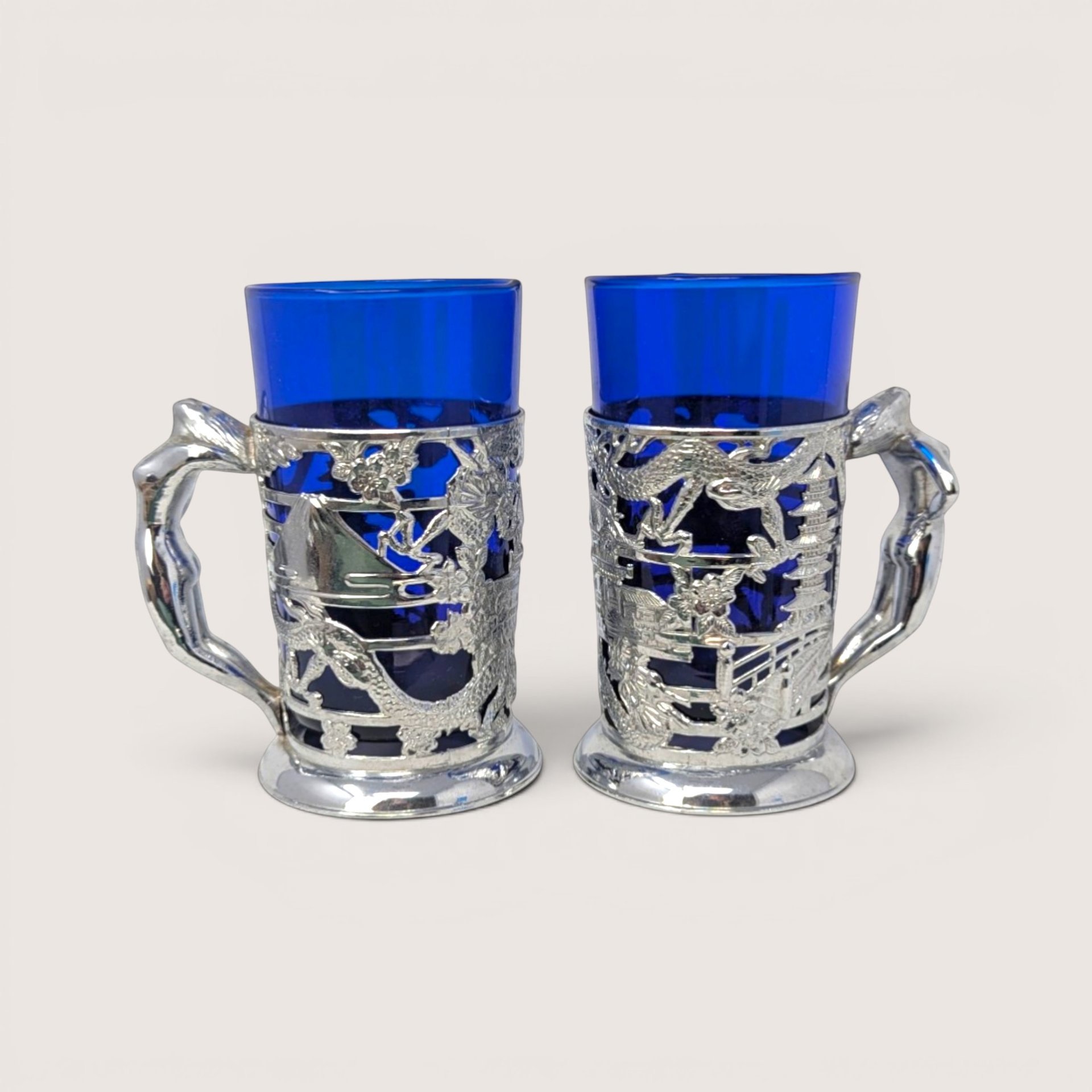 Pair of vintage silver plated metal dragon wrap tankards with cobalt blue glass inserts on a solid background.