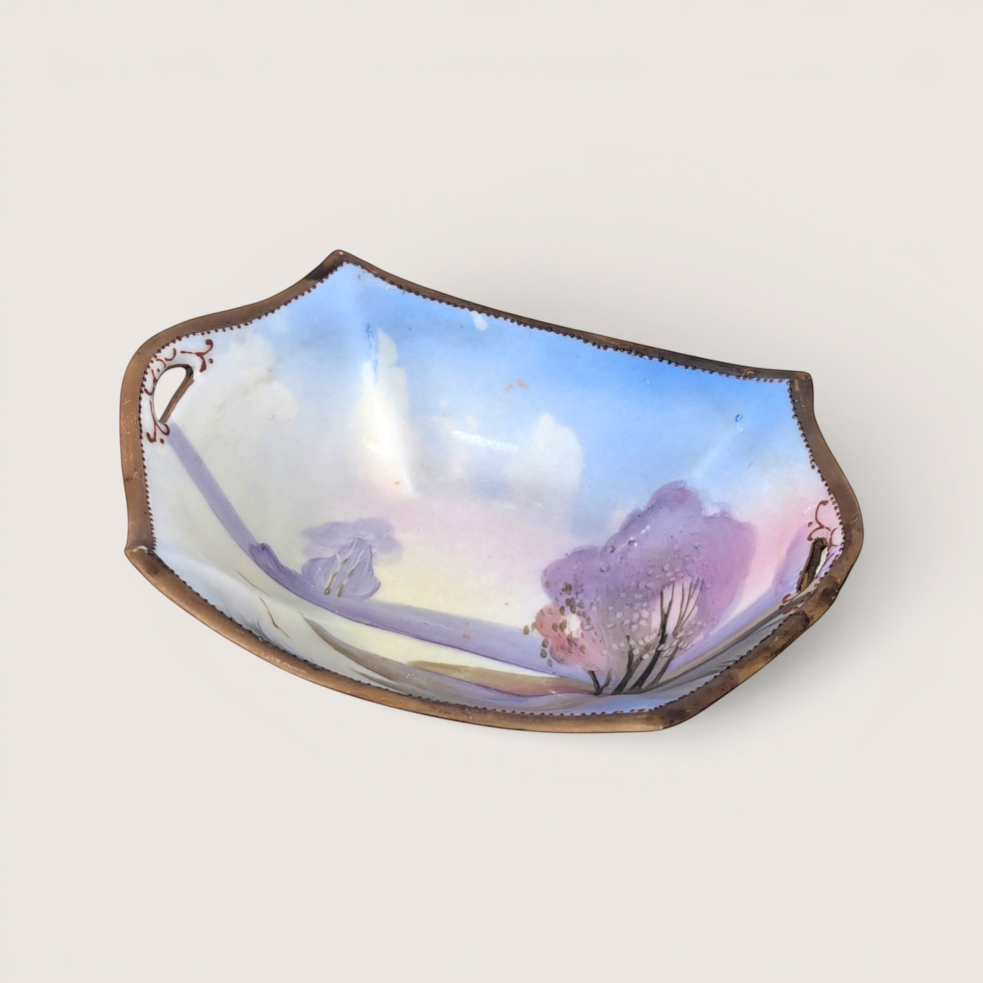 Hand painted antique Nippon porcelain dish depicting a pastel landscape with purple trees and a gold beaded rim.