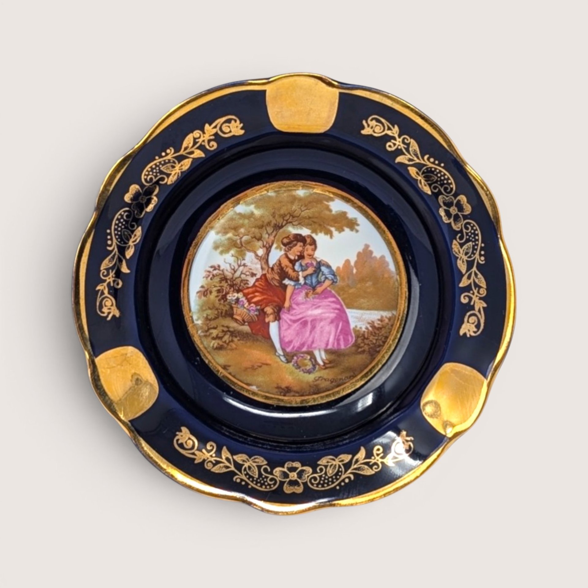 A round cobalt blue porcelain ashtray with gold scrolling trim and a central romantic couple scene on a white background