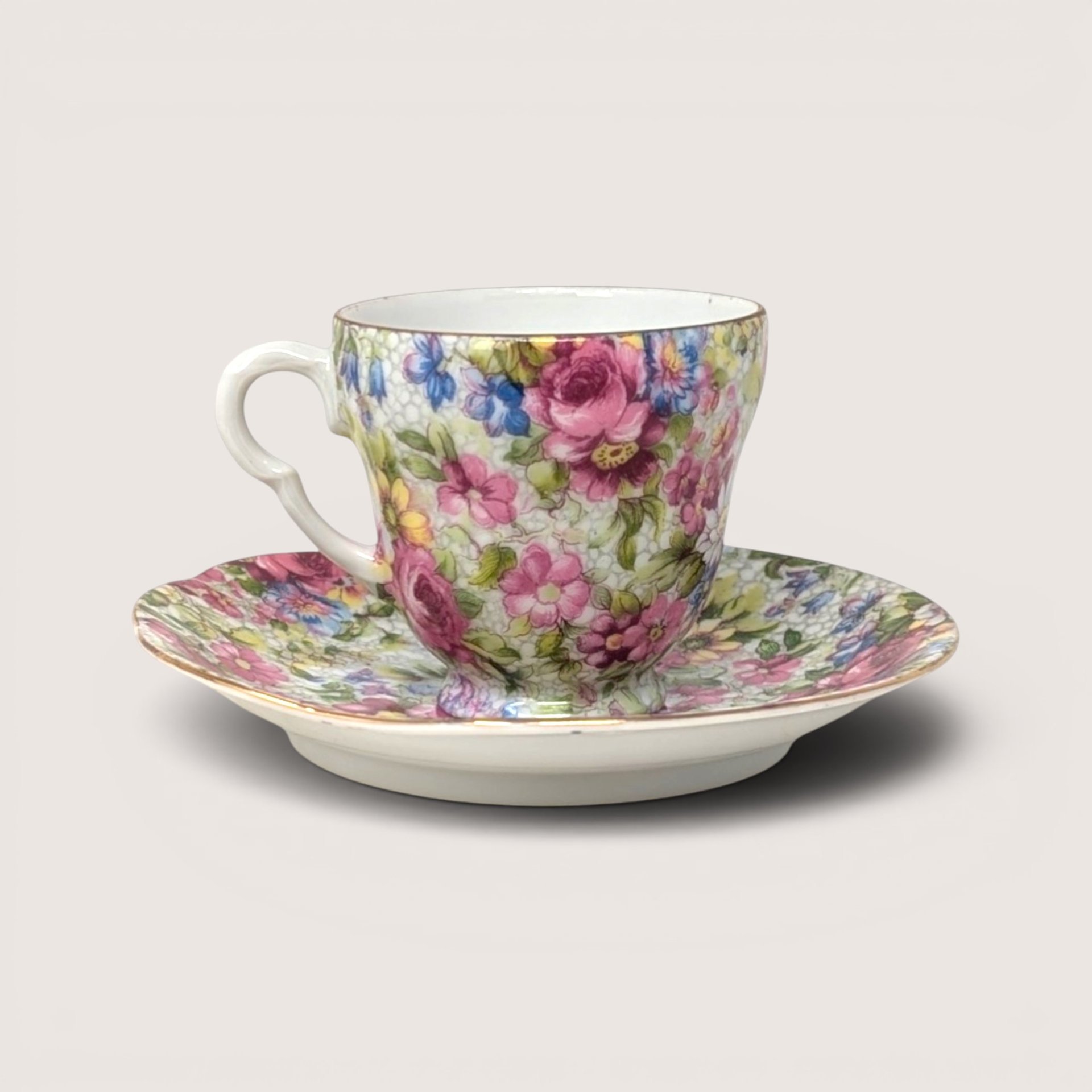 A chintz style porcelain demitasse cup and saucer with an allover pink and blue floral pattern on a white background
