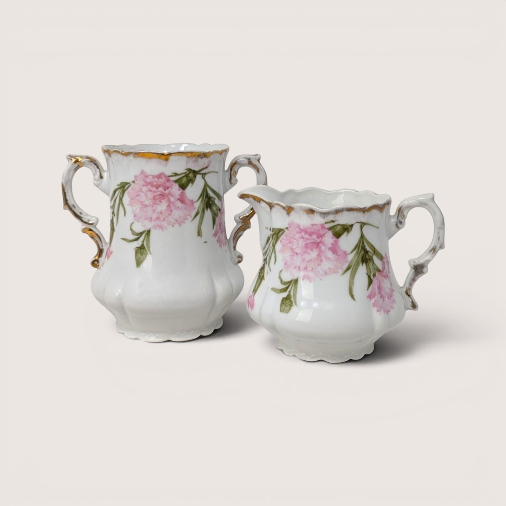A white porcelain open sugar bowl and creamer set with pink carnation blossoms and gold trim on a plain background