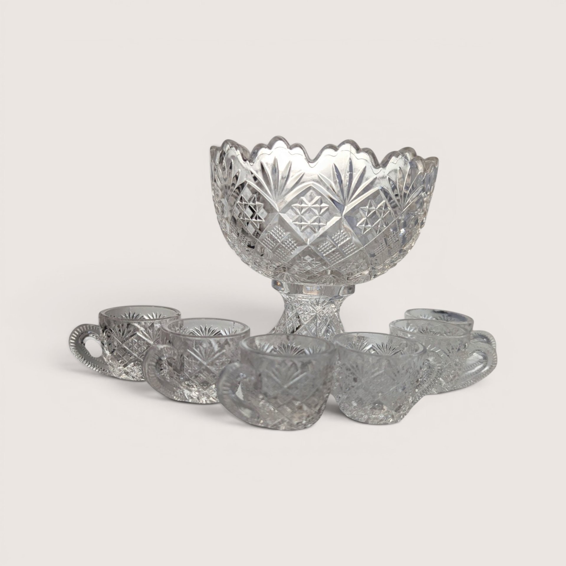 An antique clear pressed glass miniature punch bowl on a pedestal with six matching tiny cups on a plain background