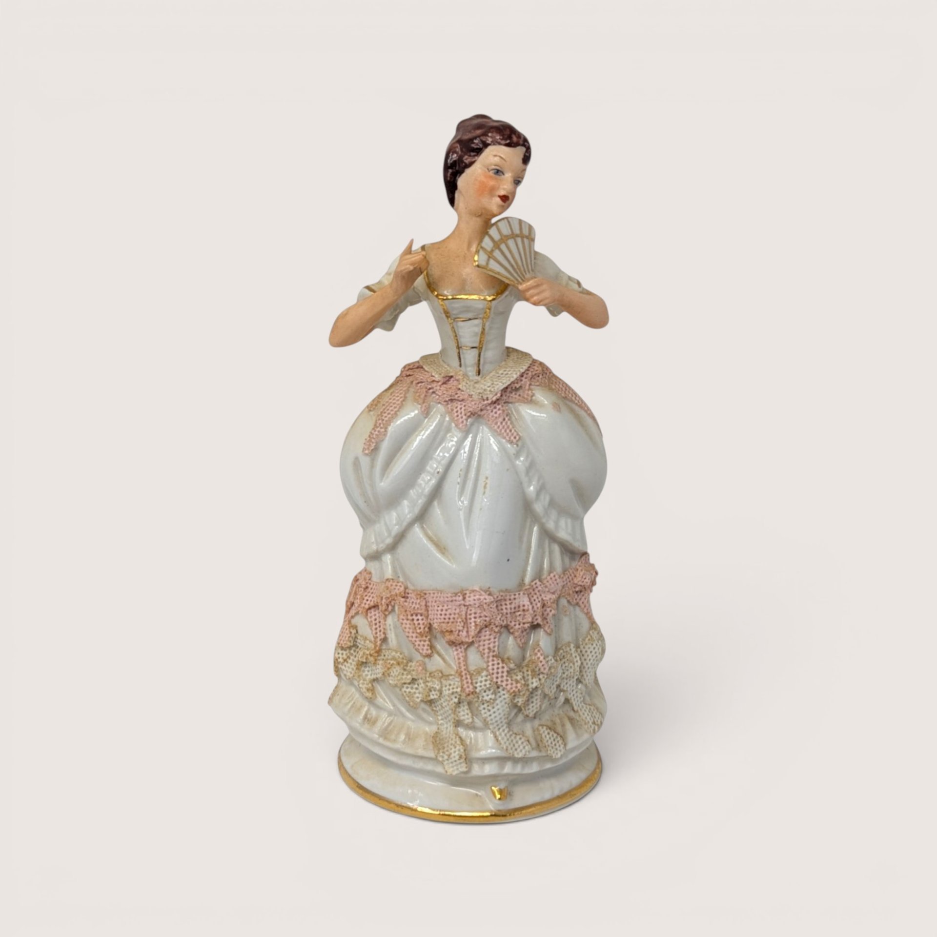 An antique porcelain figurine of a lady holding a hand fan with pink lace style details on a plain background
