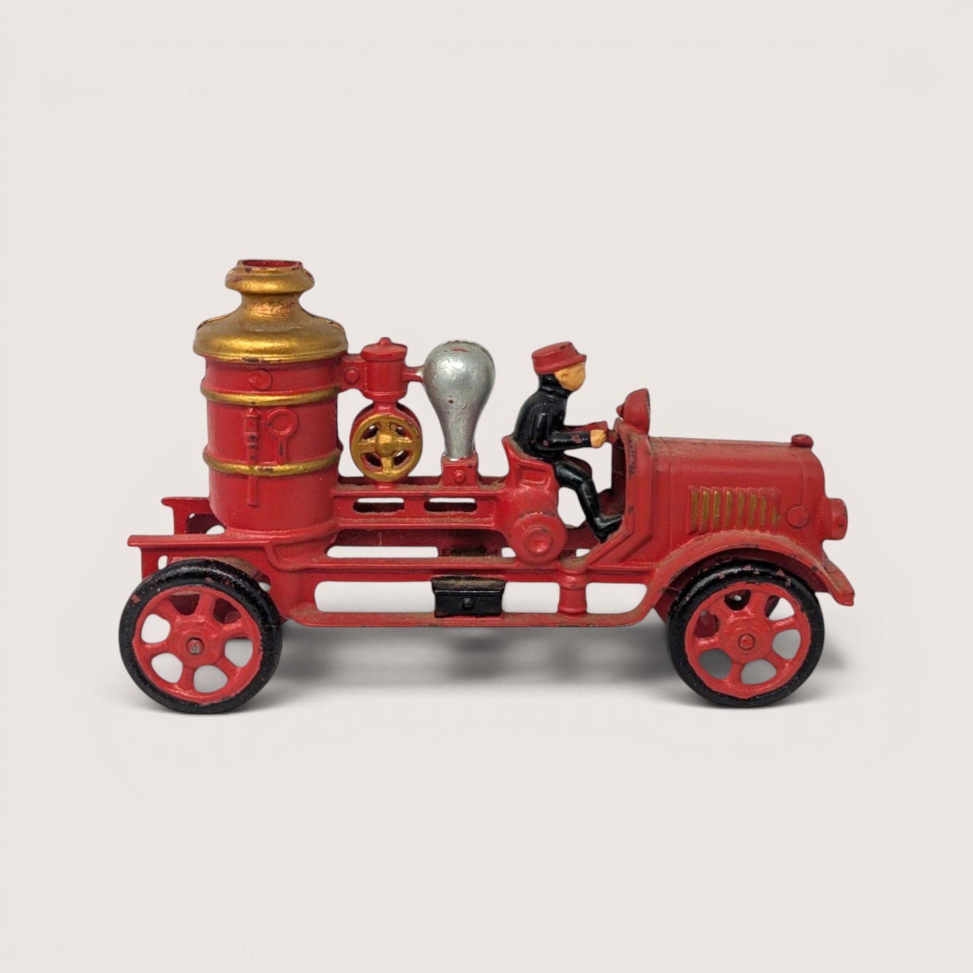 A vintage red cast iron fire pumper toy truck with spoken wheels and a small driver figure in a black uniform.