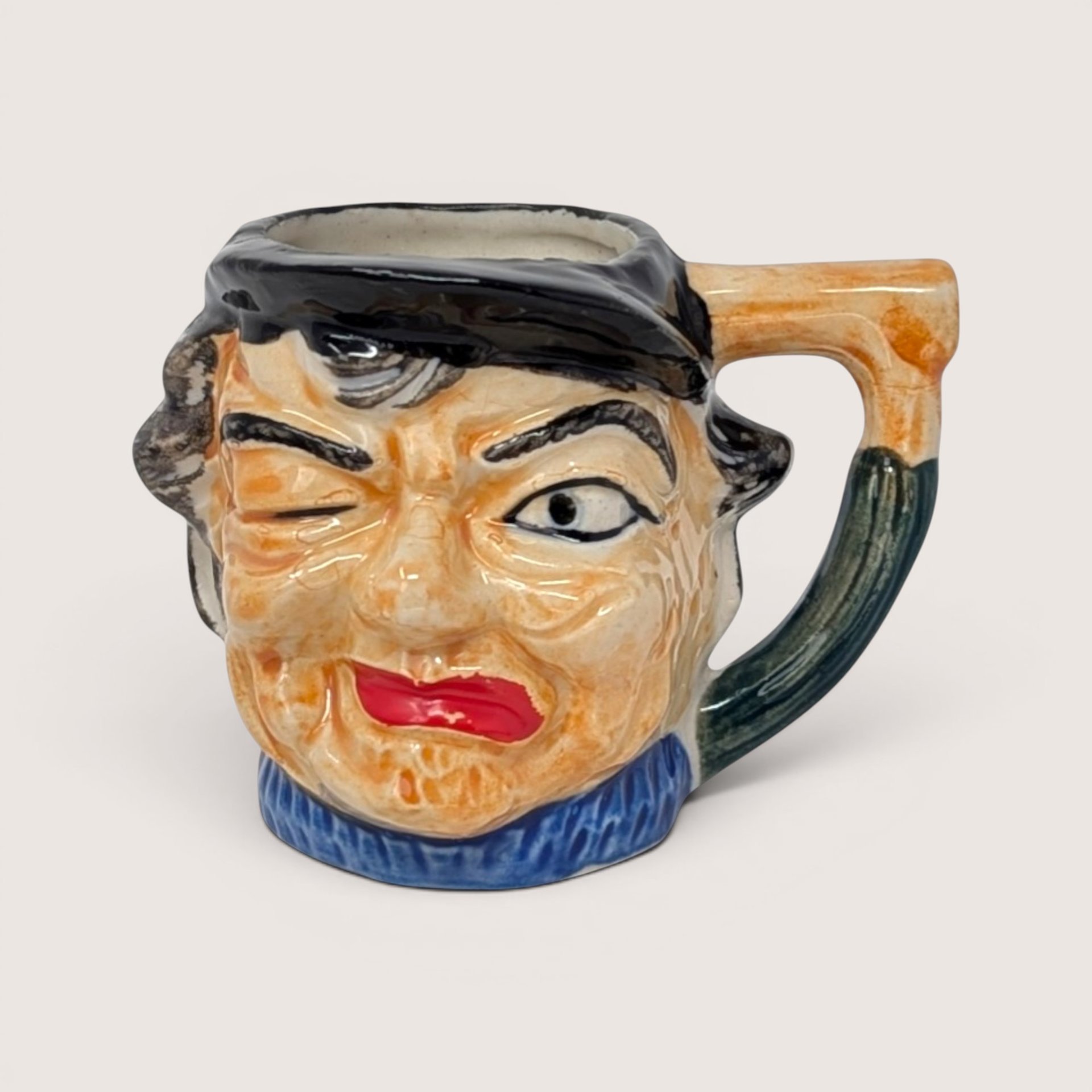 A vintage hand painted ceramic character mug depicting a winking face with a black cap and cane shaped handle