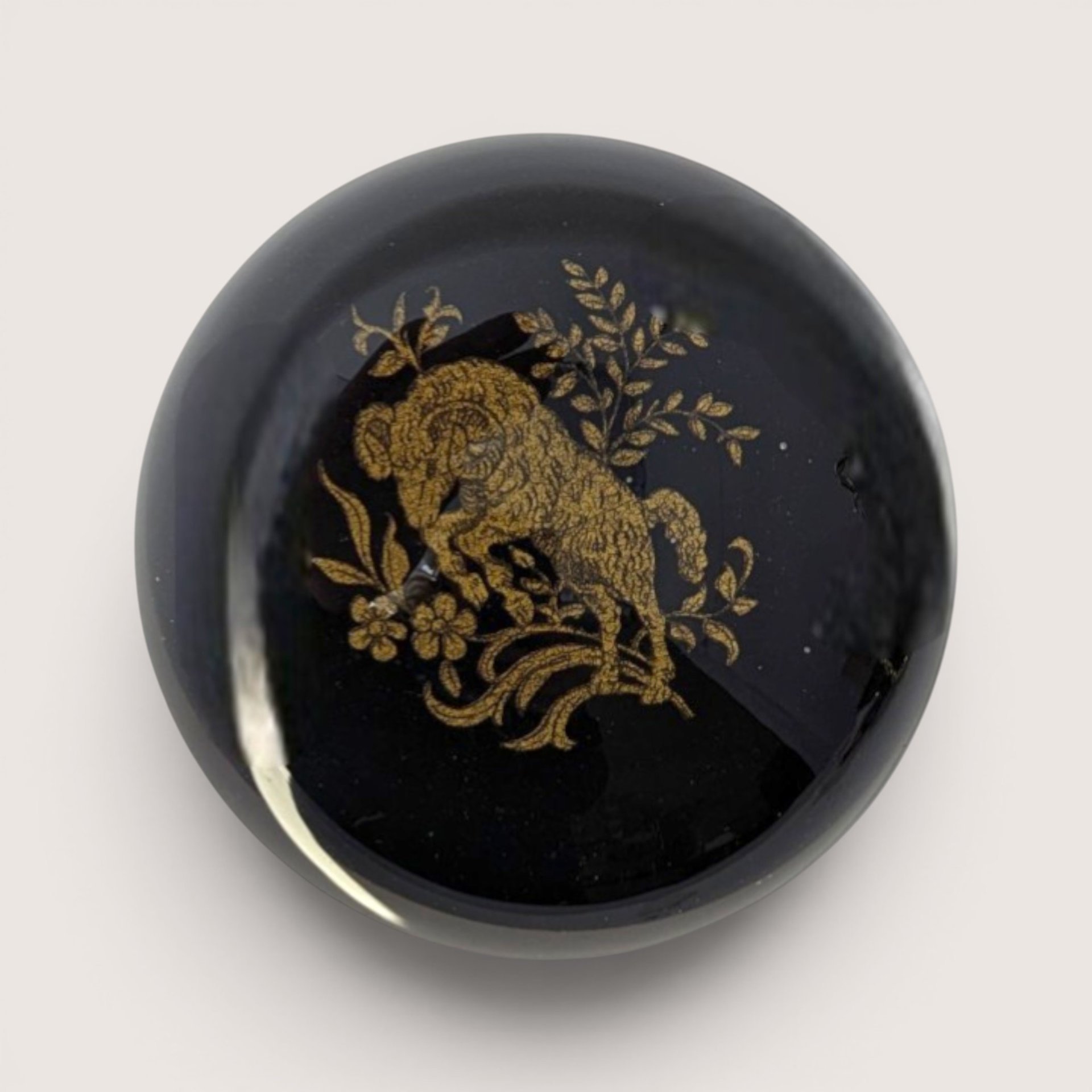 A round dome glass paperweight featuring a detailed gold ram motif over a glossy black lacquer background