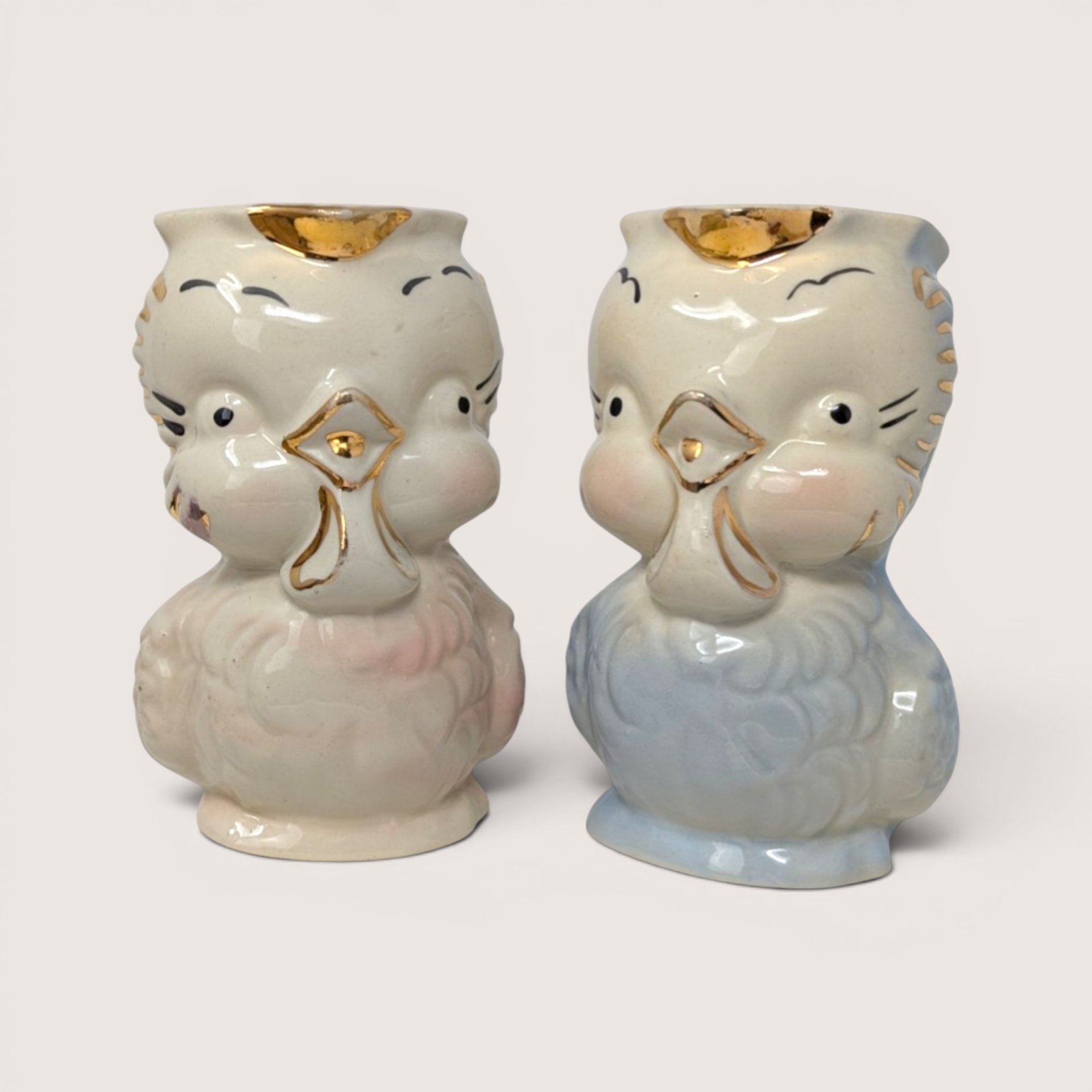A pair of vintage ceramic owl creamers in pastel pink and blue with gold accents standing side by side