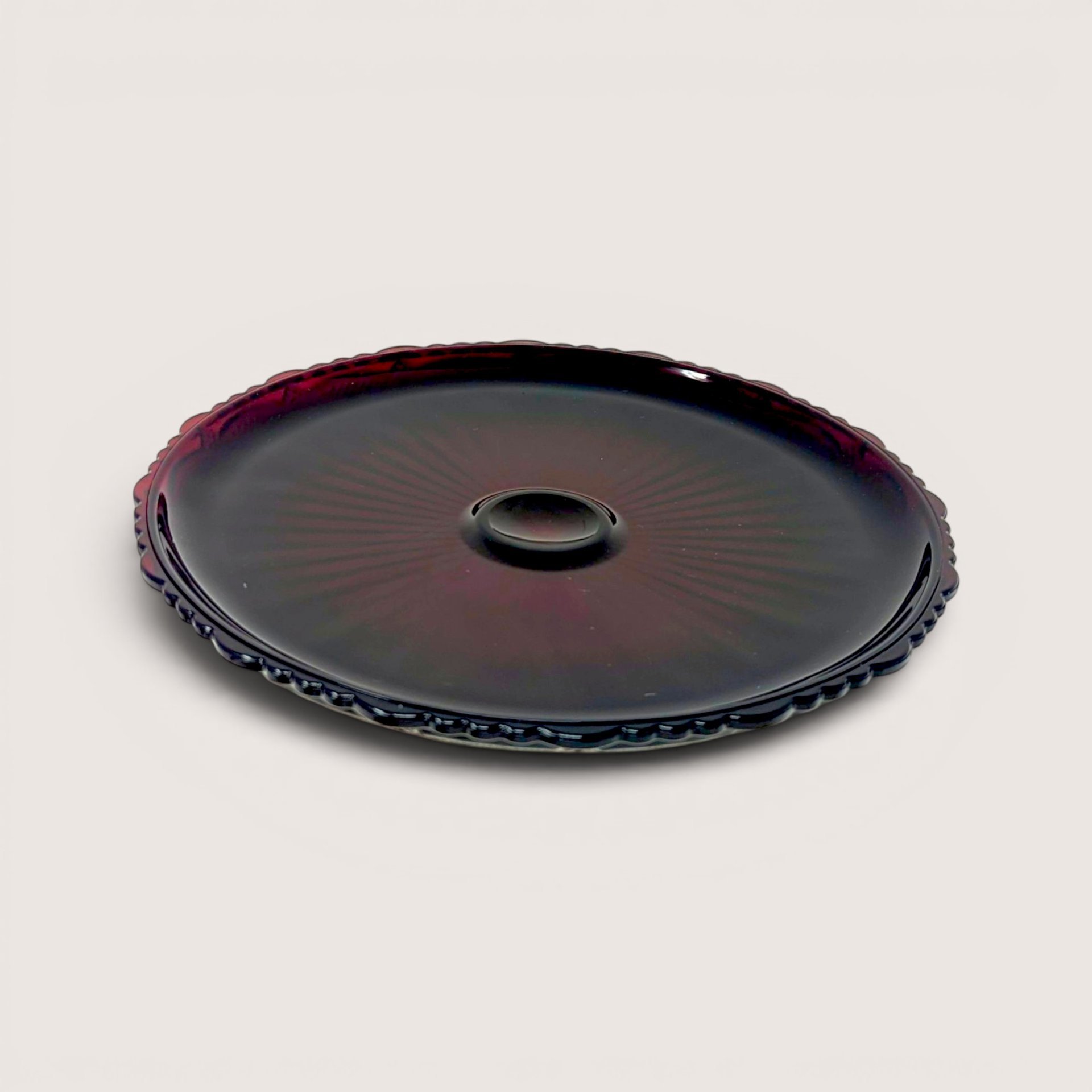 A round ruby red pressed glass cake server plate with a decorative scalloped edge on a plain background