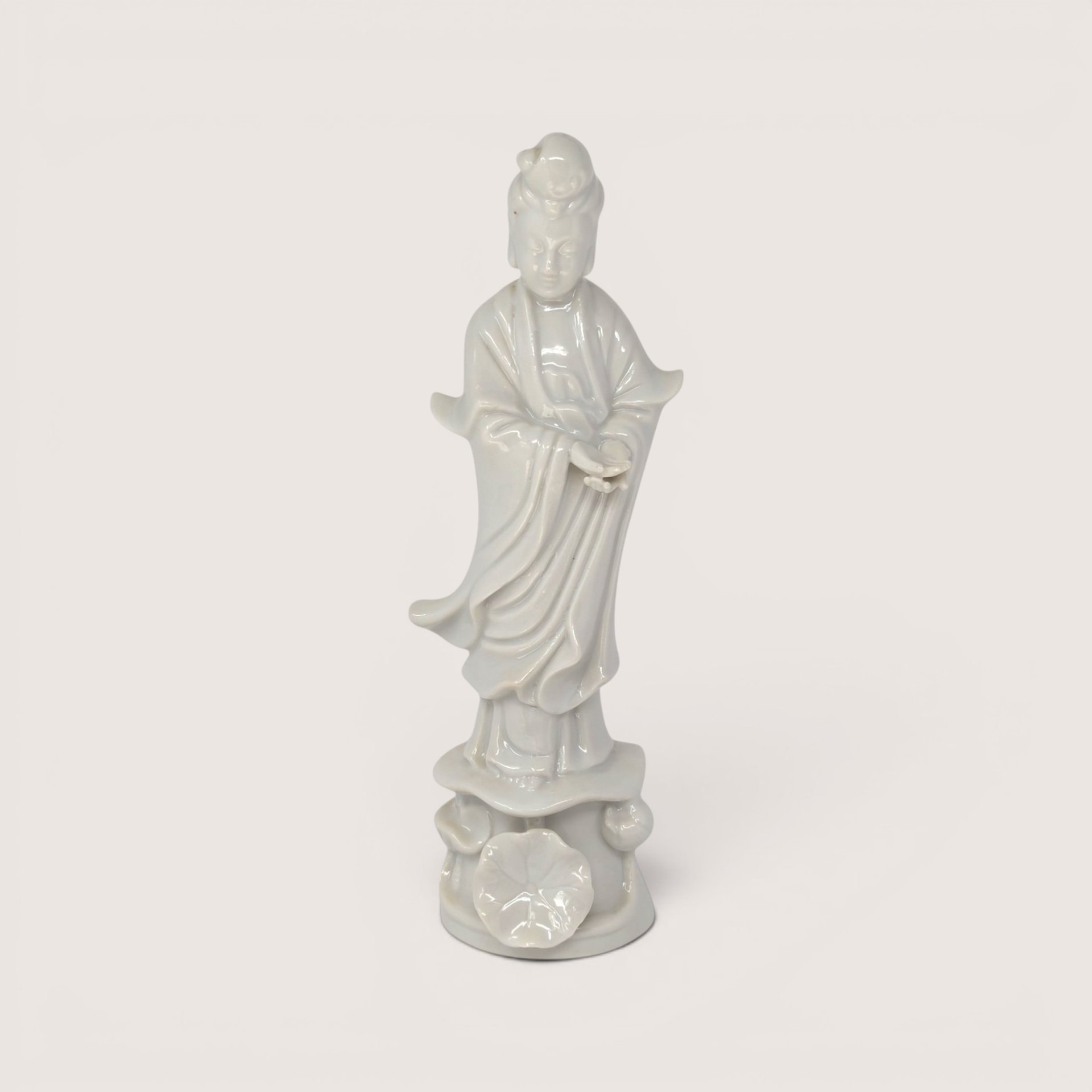 A vintage white blanc de chine porcelain Guan Yin figurine standing on a lotus base against a plain studio backdrop