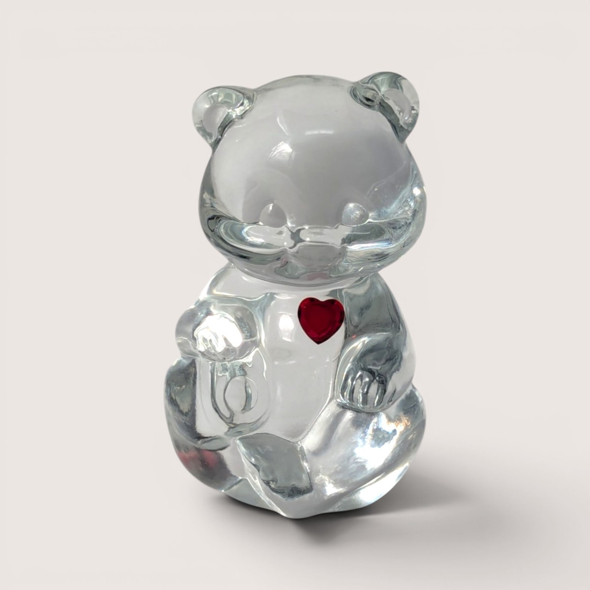 A clear glass seated teddy bear figurine featuring a small red rhinestone heart on its chest centered in the frame.