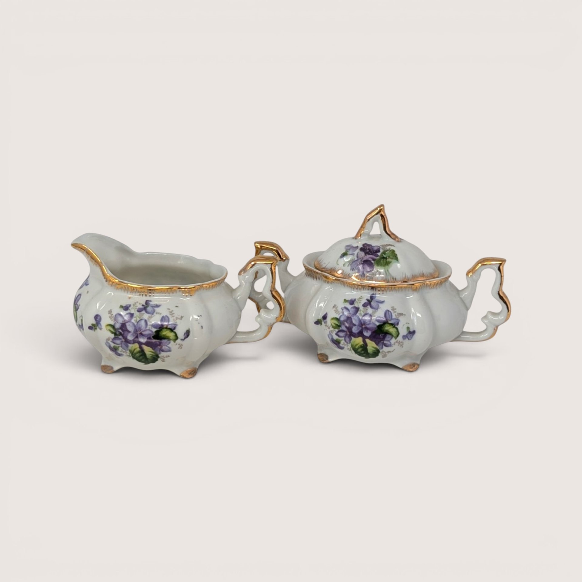 A vintage porcelain creamer and lidded sugar bowl set featuring delicate purple violet bouquets and gold trim.