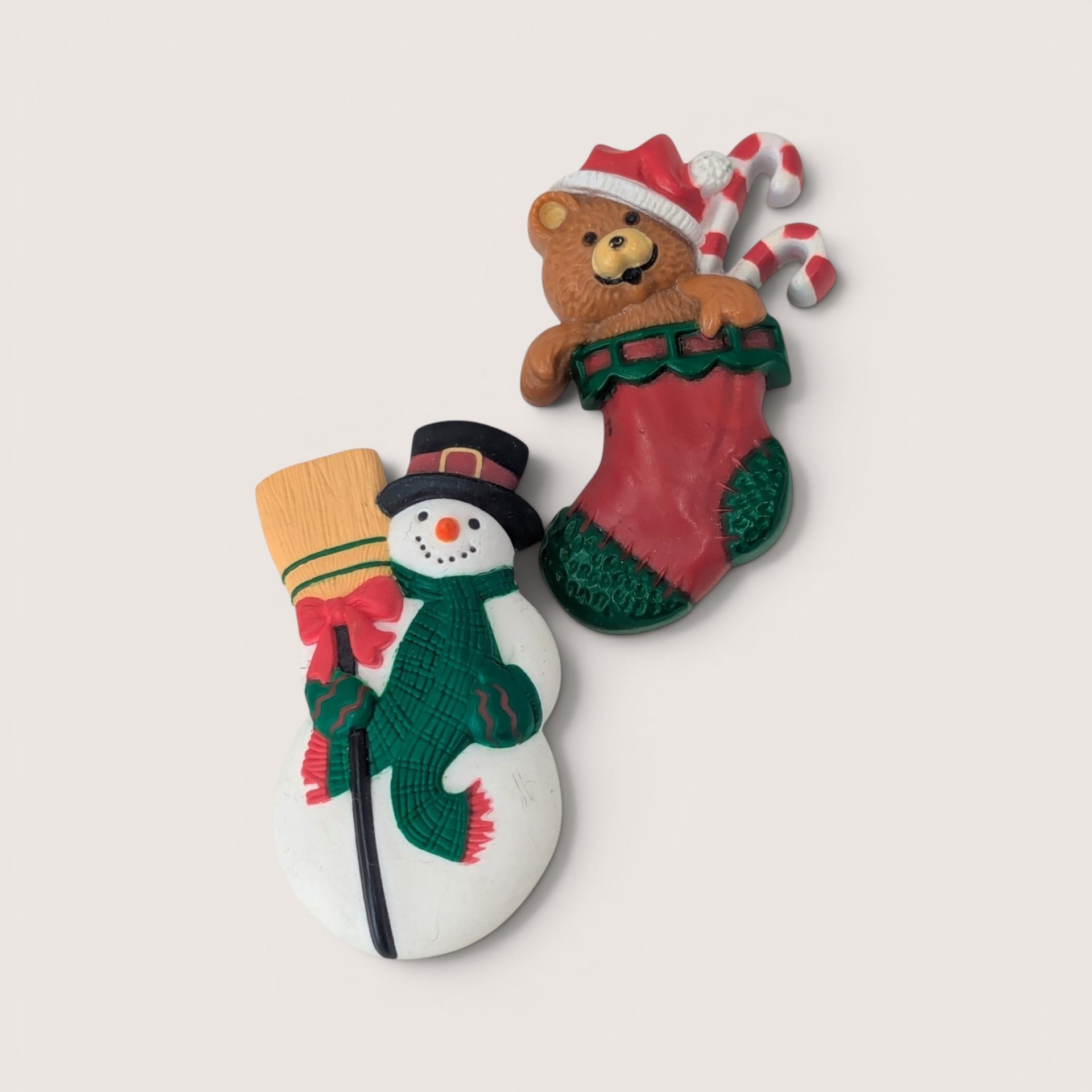 Pair of vintage Hallmark holiday brooches featuring a snowman with a broom and a teddy bear in a Christmas stocking.