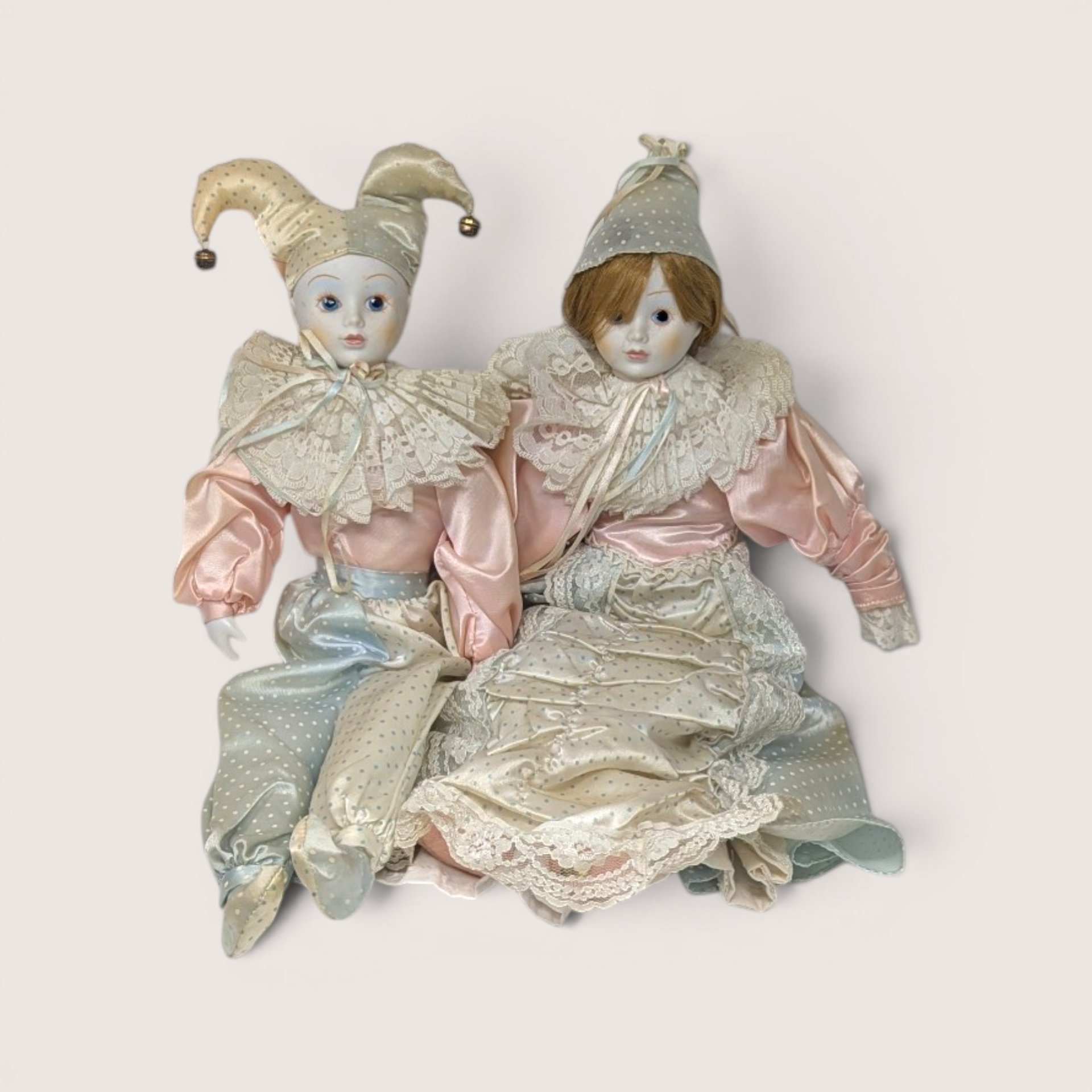 Pair of vintage porcelain Harlequin and Pierrot dolls in pastel pink, blue, and cream satin costumes with lace collars.