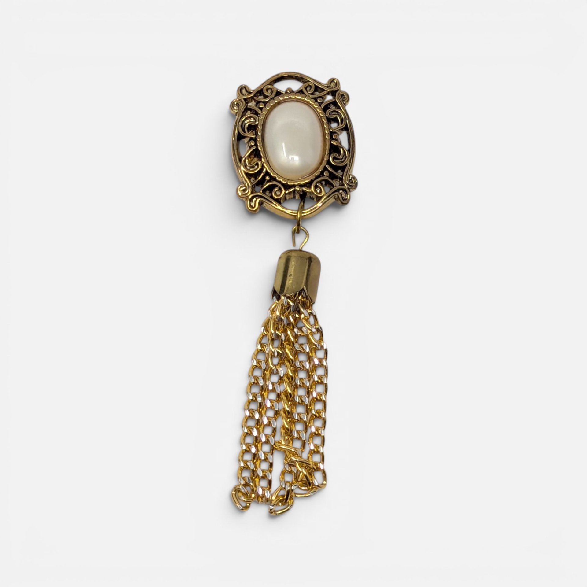Vintage gold filigree button cover with white cabochon and dangling gold chain tassel on plain background.