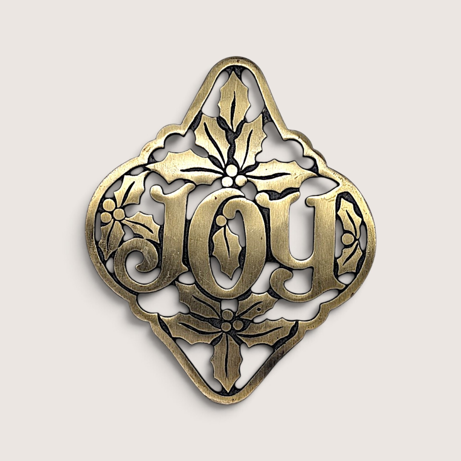 Vintage cast brass holiday trivet featuring openwork "JOY" typography framed by sculpted holly leaves and berries.