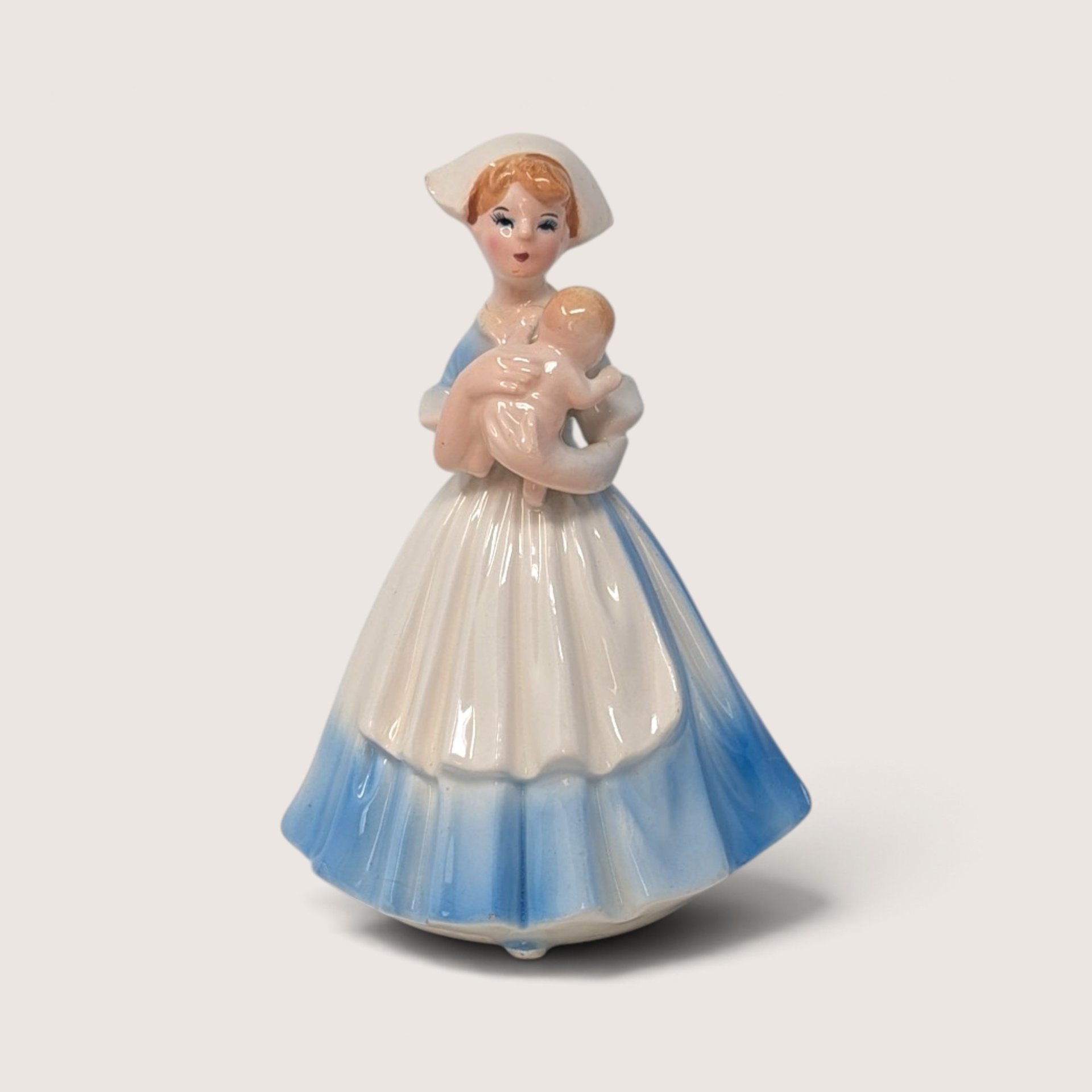 Vintage porcelain figurine of a nurse in a blue and white dress holding a baby, with a close-up highlighting a small chip on her hat.