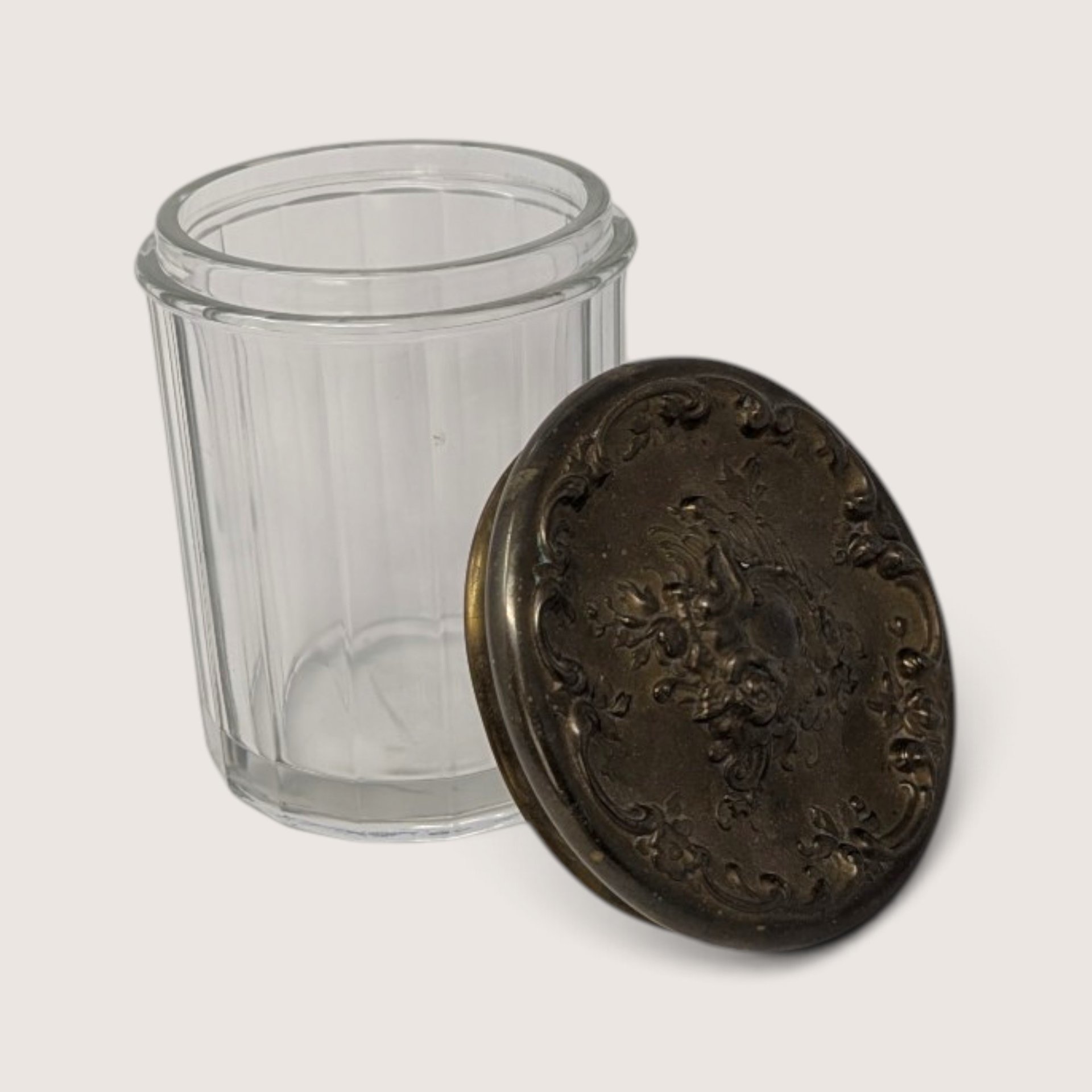 Vintage ribbed glass jar featuring an ornate, heavily embossed metal lid resting on top.