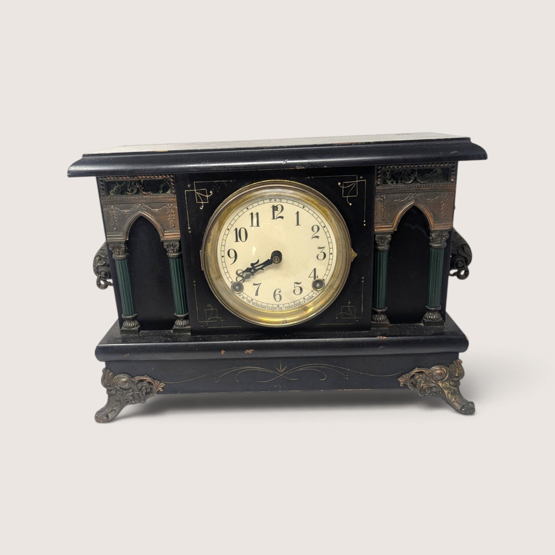 Front view of the antique black architectural mantel clock showing the detailed dial with arabic numerals, ornate side pillars, and decorative cast metal feet.   