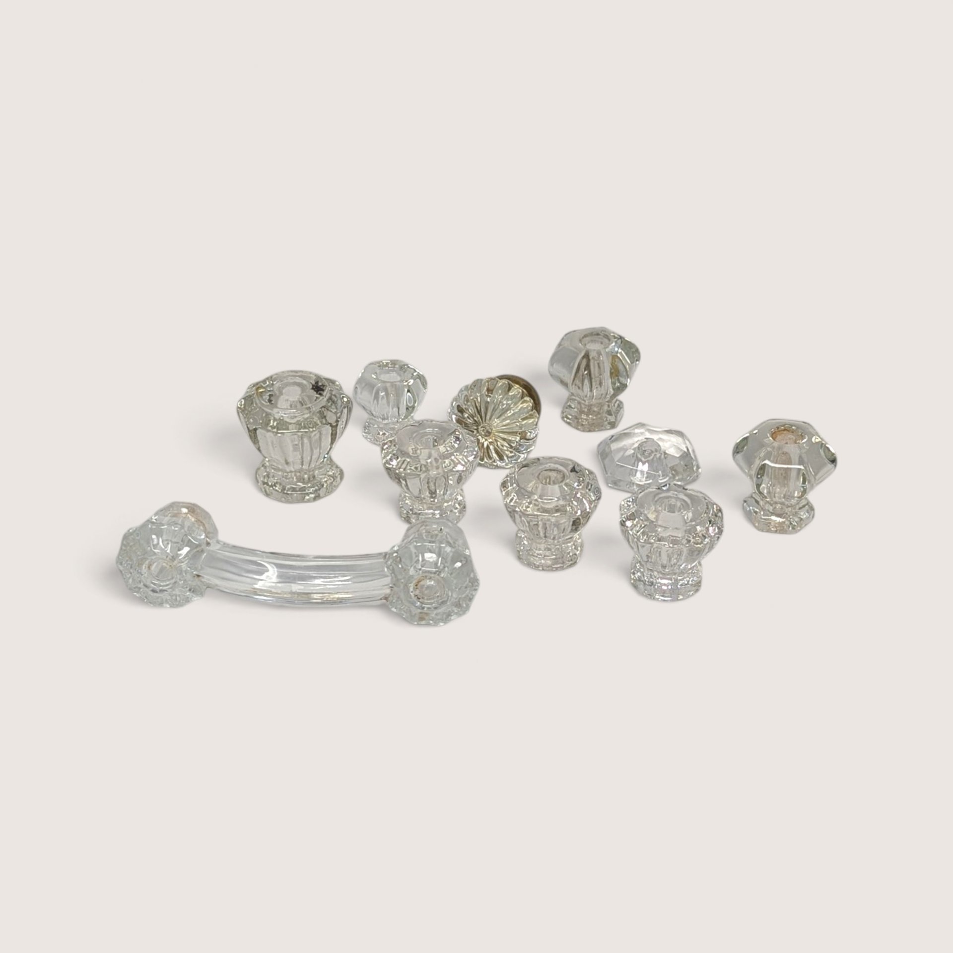 A collection of 10 assorted vintage clear glass cabinet knobs and one curved drawer pull laid out on a neutral background.