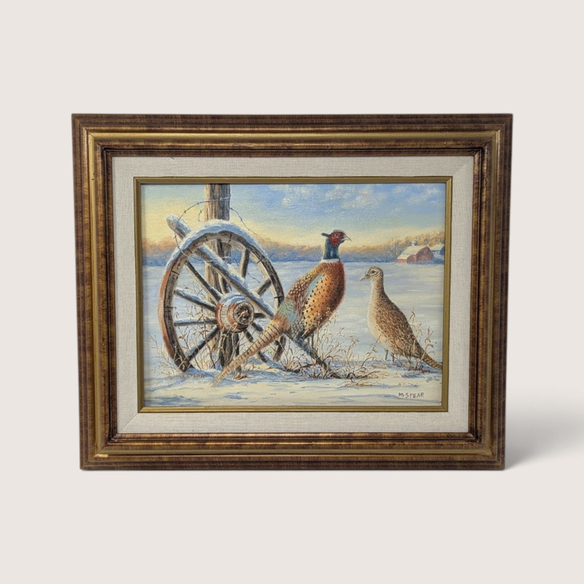 An original framed painting by Martha Spear depicting a rooster and hen pheasant standing in a snowy field beside an old wooden wagon wheel, set inside a textured wood frame with a fabric liner, showing the Fredrix canvas backing on the rear.